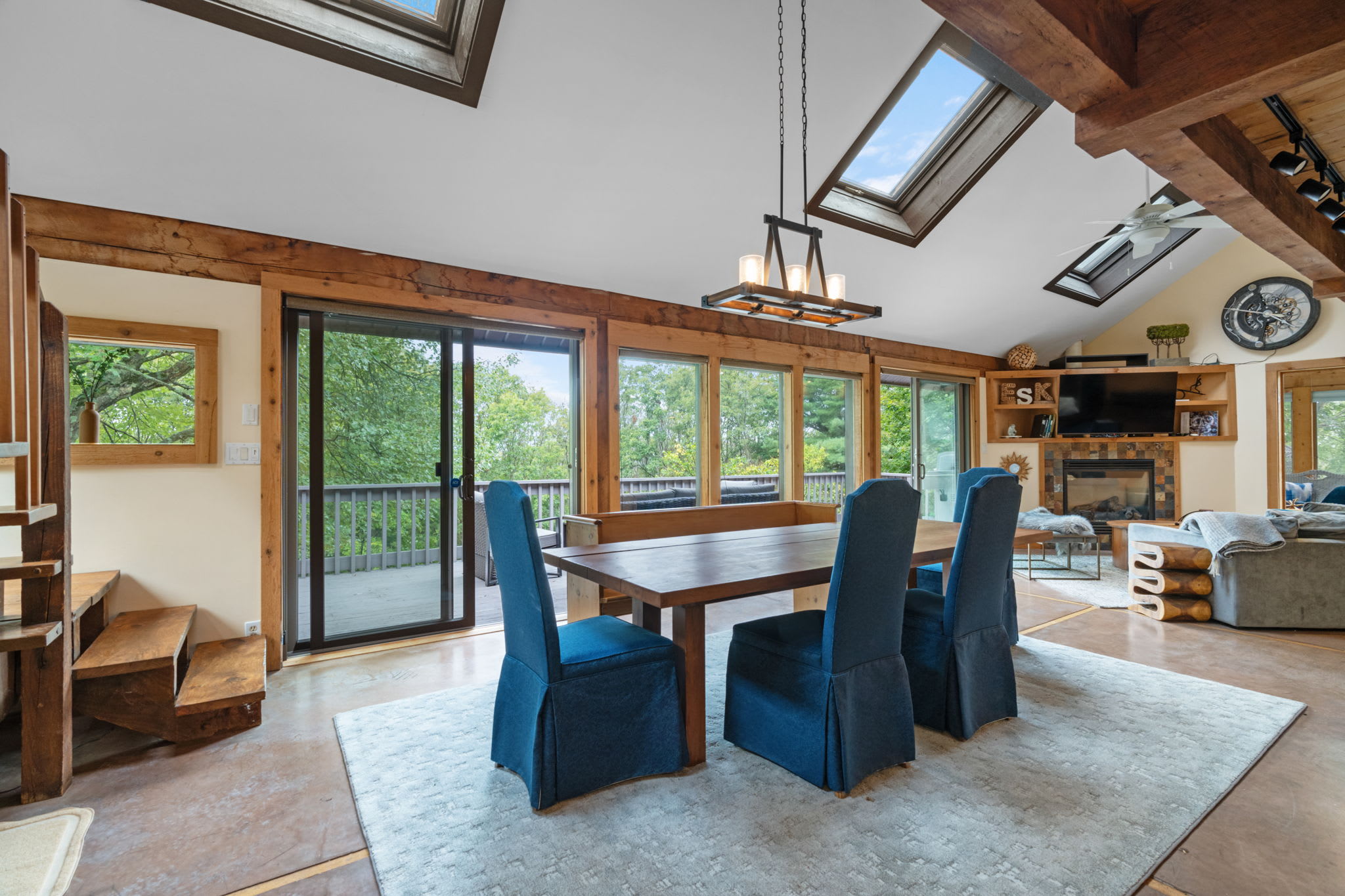 Bright dining area with a long wooden table and blue upholstered chairs, featuring multiple skylights, exposed wooden beams, and sliding glass doors leading to an outdoor deck.