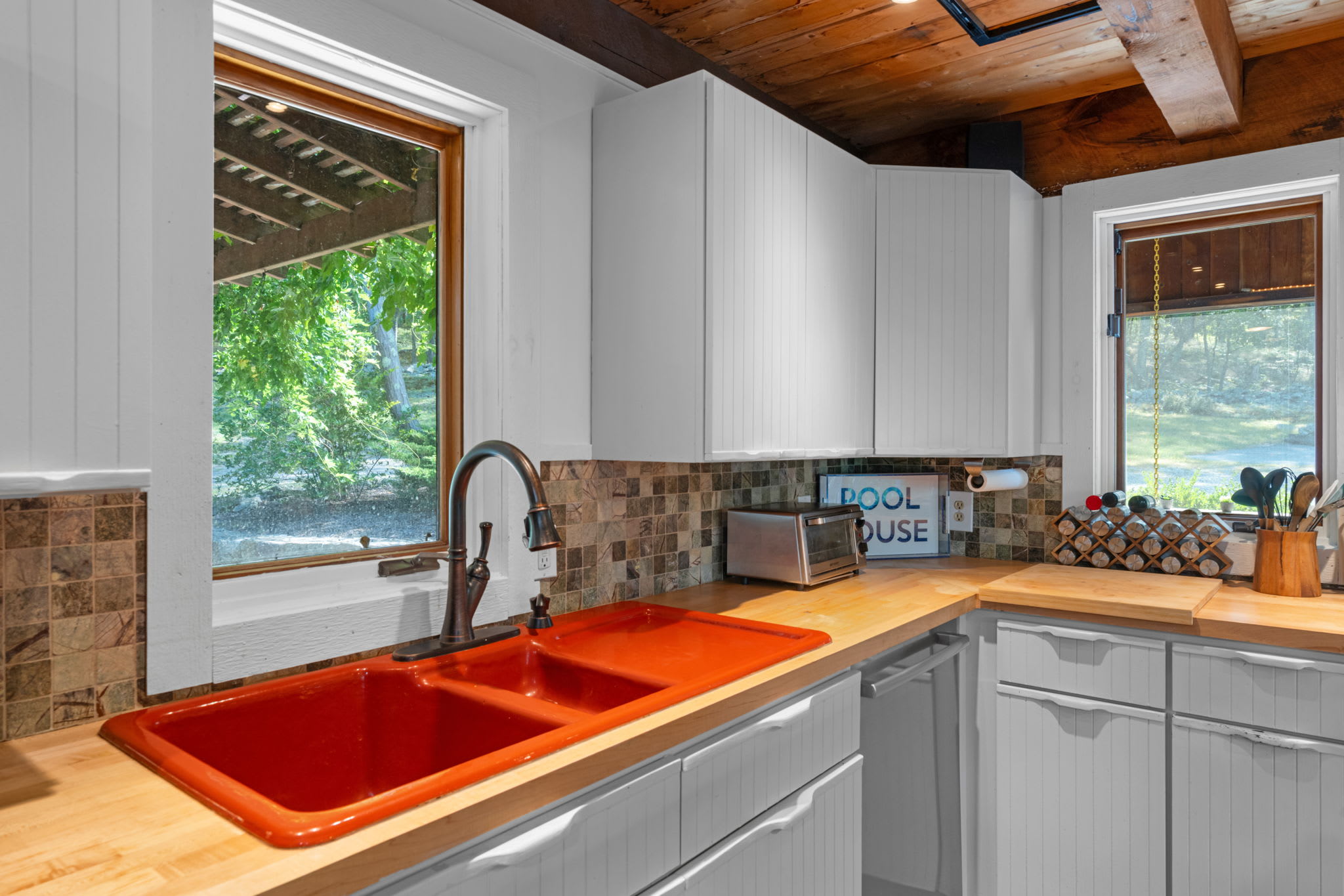 A close-up of the kitchen sink area featuring a vibrant red double basin sink set into a butcher block countertop, with a mosaic tile backsplash and white shaker cabinets above.