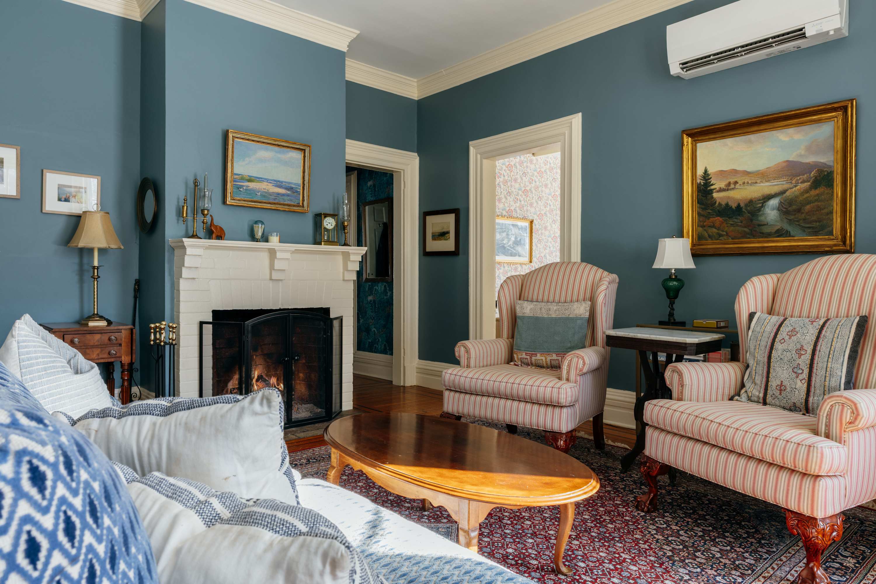 A comfortable living room with blue walls, a white fireplace, and a patterned area rug. Two striped armchairs and a white sofa with a blue throw blanket are arranged around a wooden coffee table.
