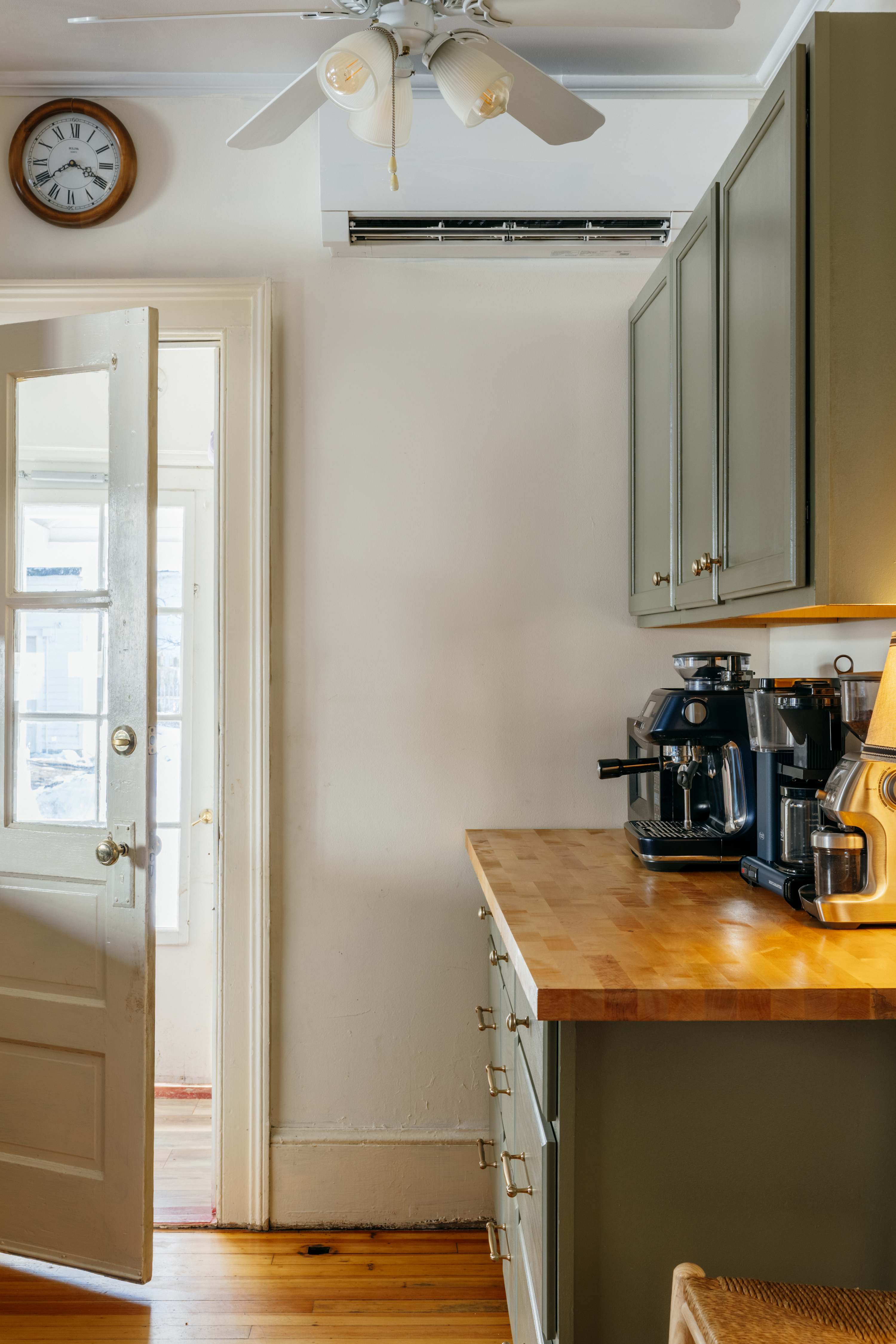 Bright kitchen featuring olive green shaker-style cabinets, a warm butcher block countertop, and modern coffee machines. A vintage ceiling fan and a wall clock add character, with a white door leading to another space.