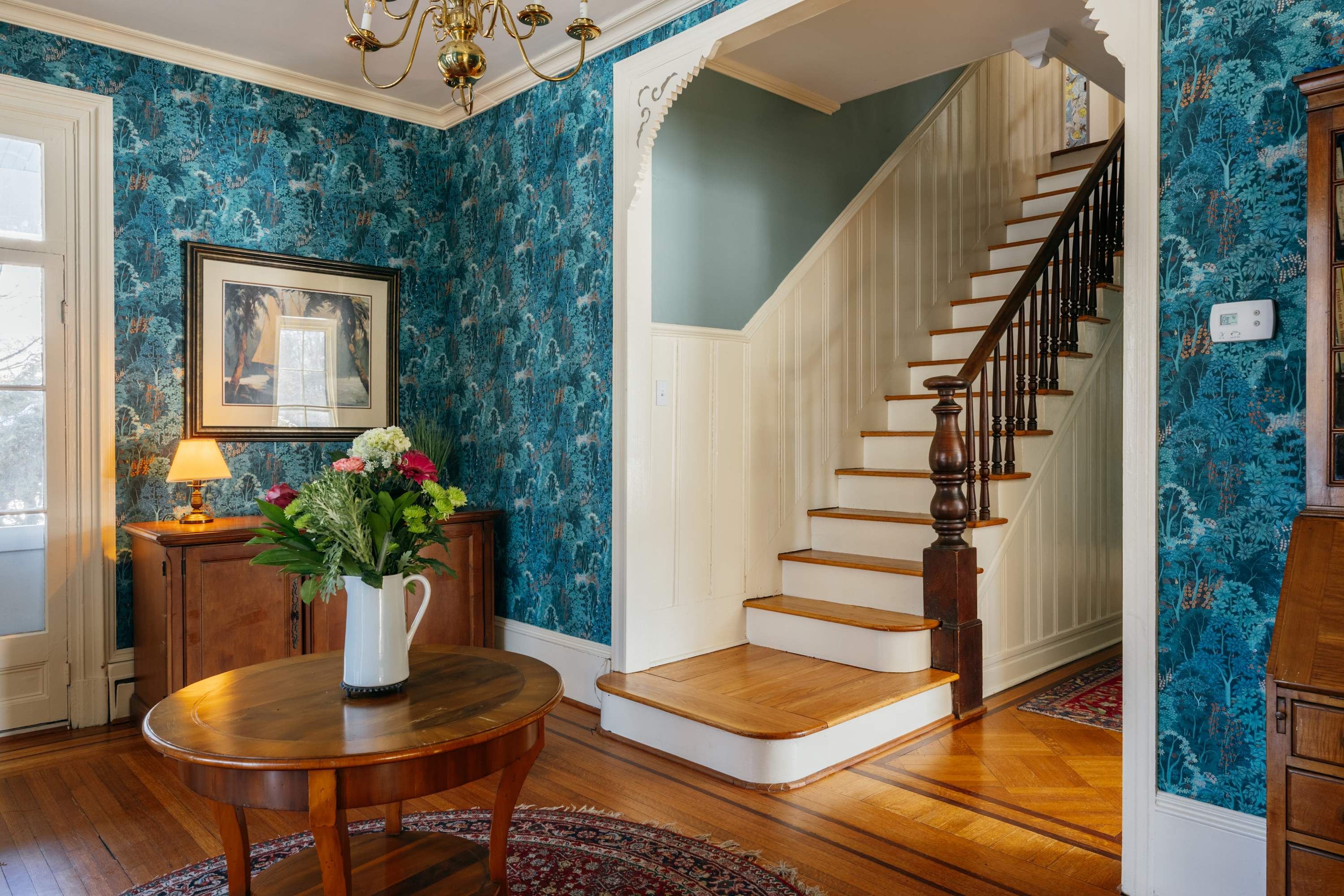 A welcoming foyer featuring vibrant teal wallpaper with a nature motif, polished hardwood floors, and a grand wooden staircase with white risers and a dark banister. A round wooden table with flowers sits on a patterned rug.