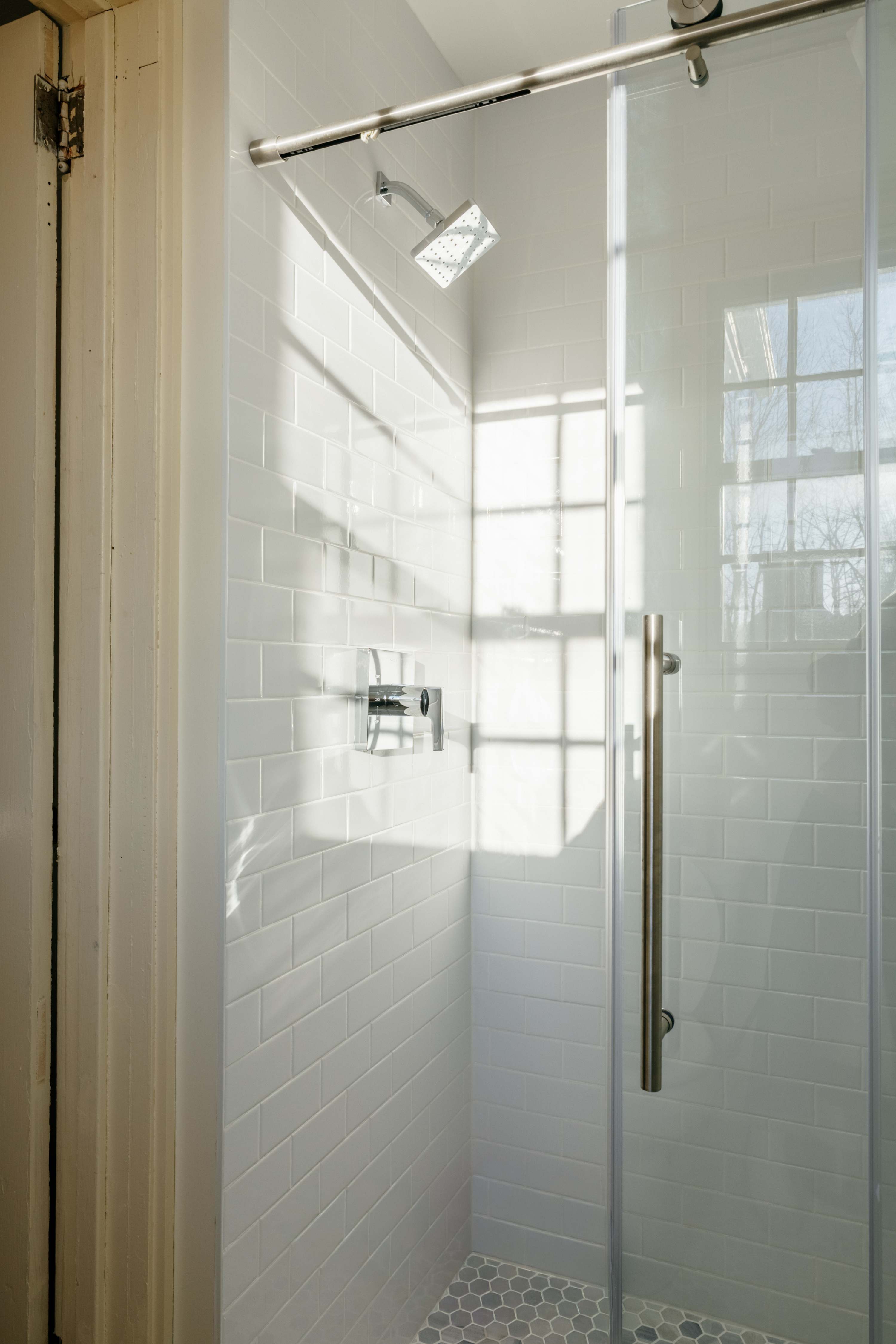 Bright shower enclosure with white subway tiles on the walls and a gray hexagonal tile floor. A modern chrome shower head and controls are visible, with sunlight streaming through a window.