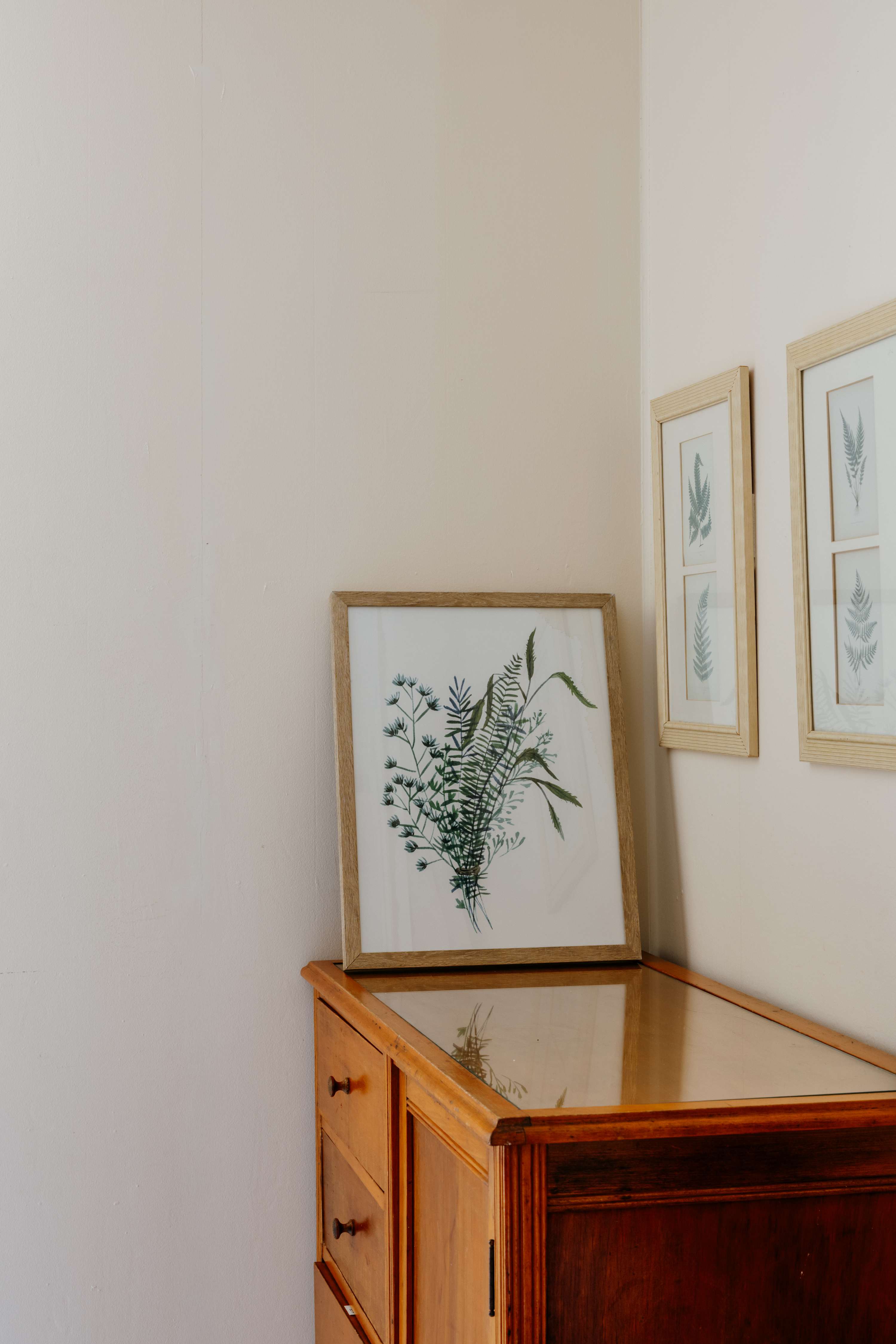 Detail shot of a wooden dresser with a glass top, displaying framed botanical prints. The walls are painted a soft, light cream color.