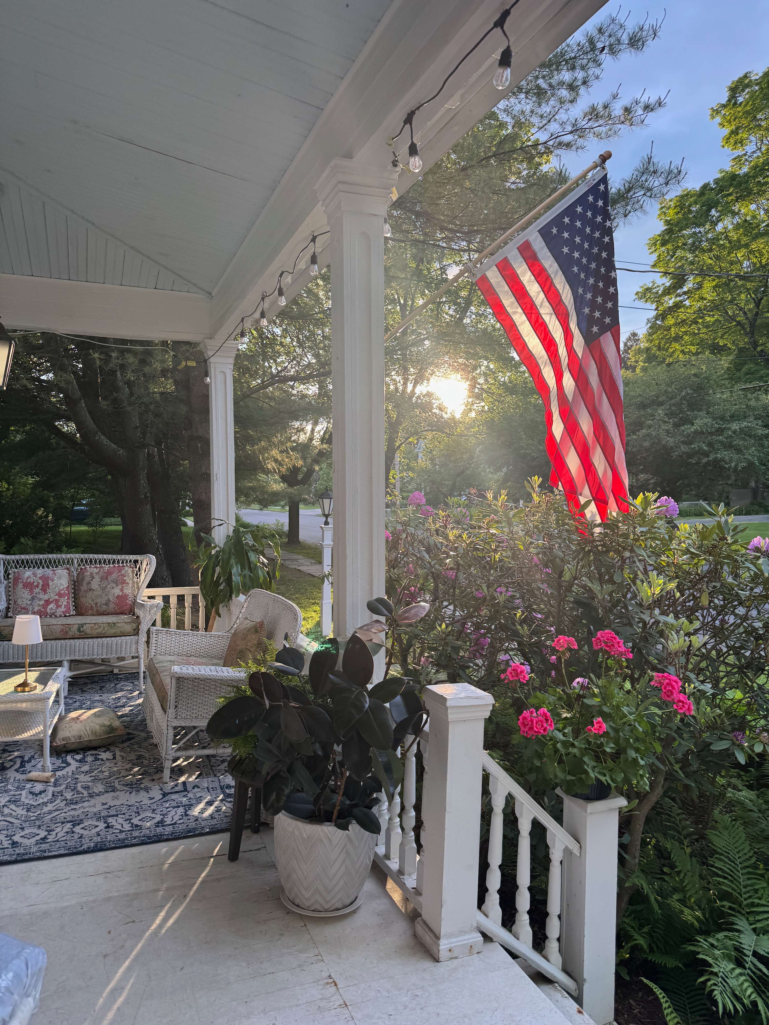 A cozy front porch with white columns and string lights, featuring wicker furniture and potted plants. An American flag hangs prominently, illuminated by the setting sun filtering through the surrounding trees.