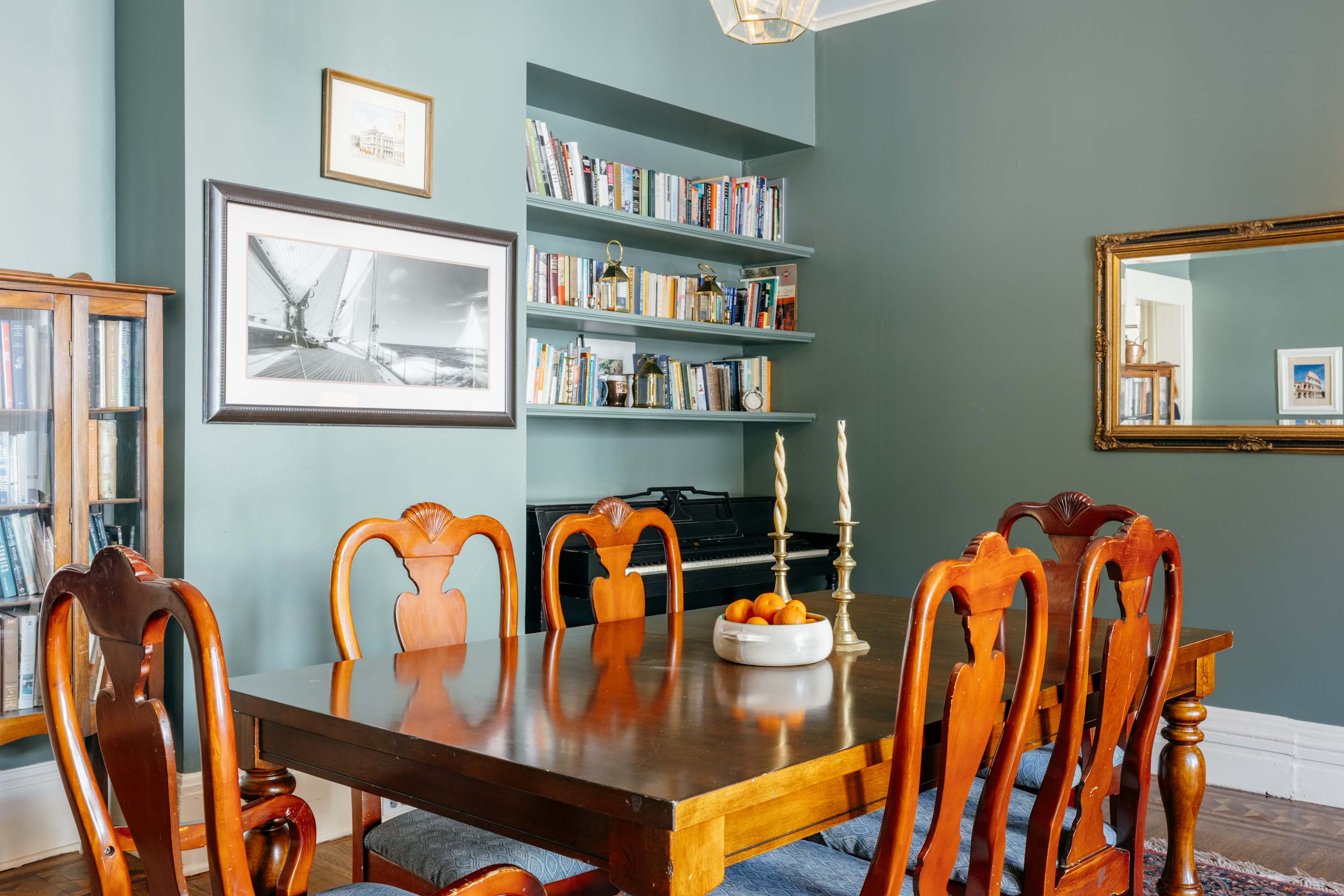 A formal dining room with sage green walls, a long wooden dining table, and six matching wooden chairs with blue upholstered seats. Built-in shelves filled with books and a piano are visible in the background.