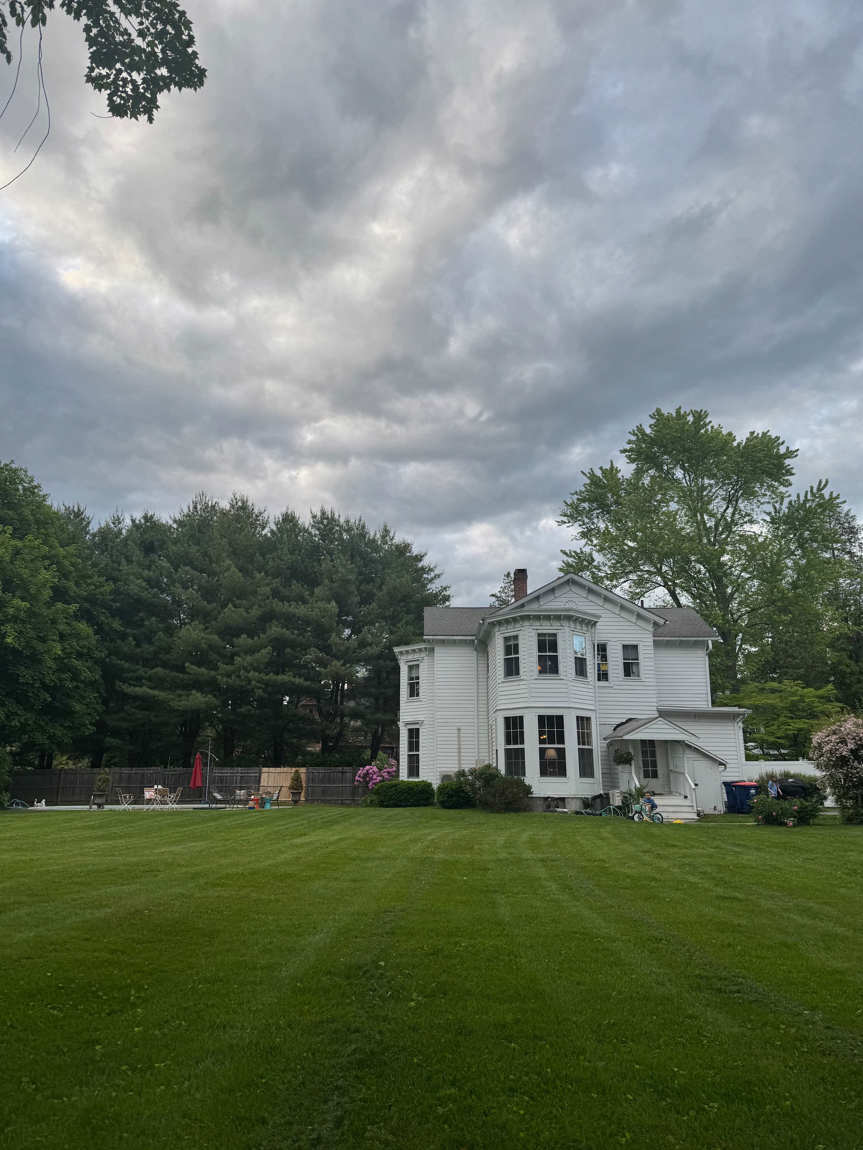 Expansive backyard featuring a large green lawn and the white exterior of the house under a cloudy sky. Mature trees line the property, with outdoor seating visible in the distance.