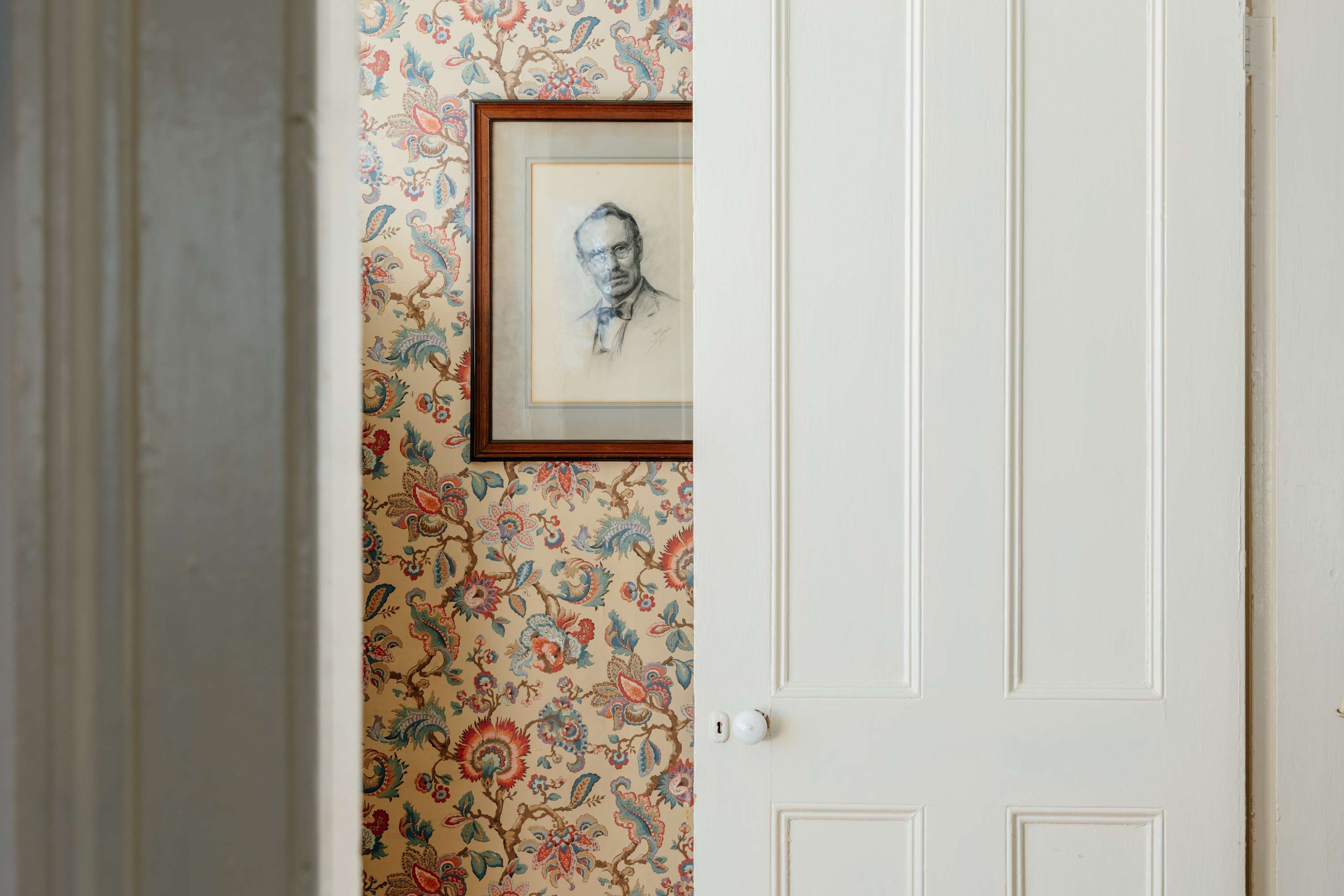 A partially open white paneled door reveals a wall adorned with vibrant floral wallpaper in blues, reds, and yellows, featuring a framed pencil portrait of a man.