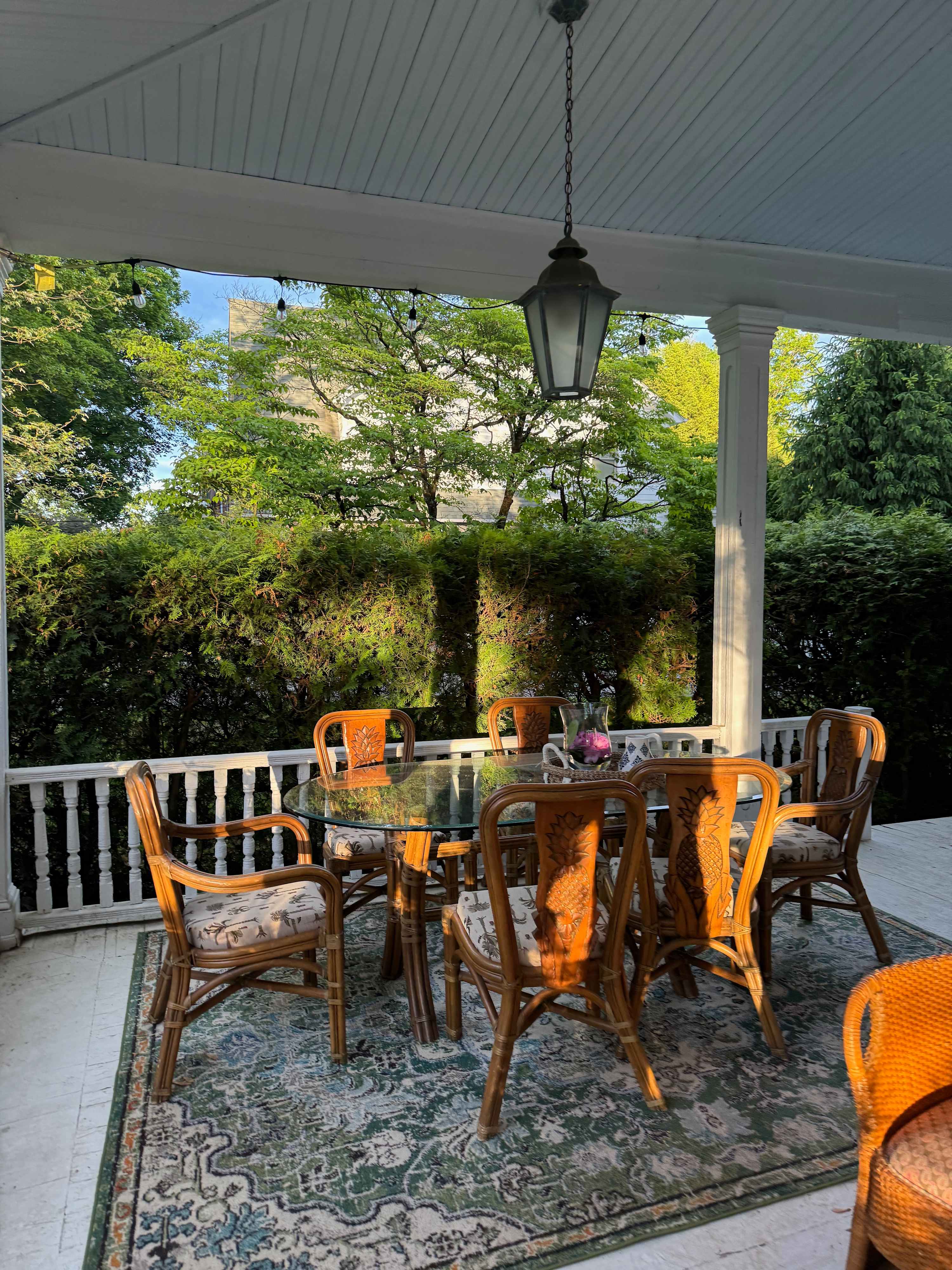 A charming outdoor porch with a glass-top dining table and wicker chairs, set on a patterned outdoor rug. Lush green foliage provides a private backdrop, and a hanging lantern illuminates the space.