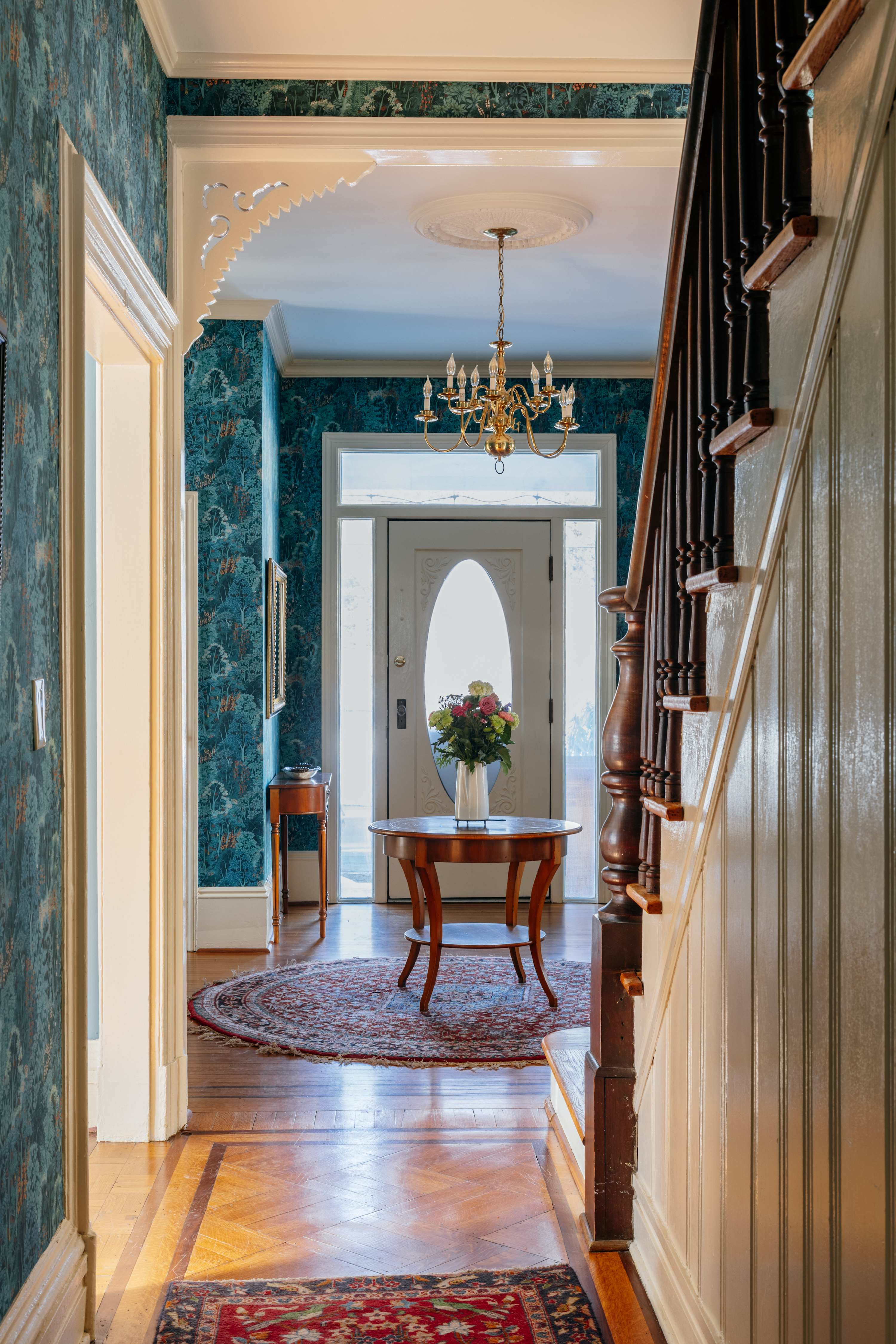 A welcoming hallway leading to the front door, featuring ornate blue patterned wallpaper, a classic wooden staircase with dark banisters, and a round table with flowers. A brass chandelier hangs from the ceiling, adding a touch of elegance.