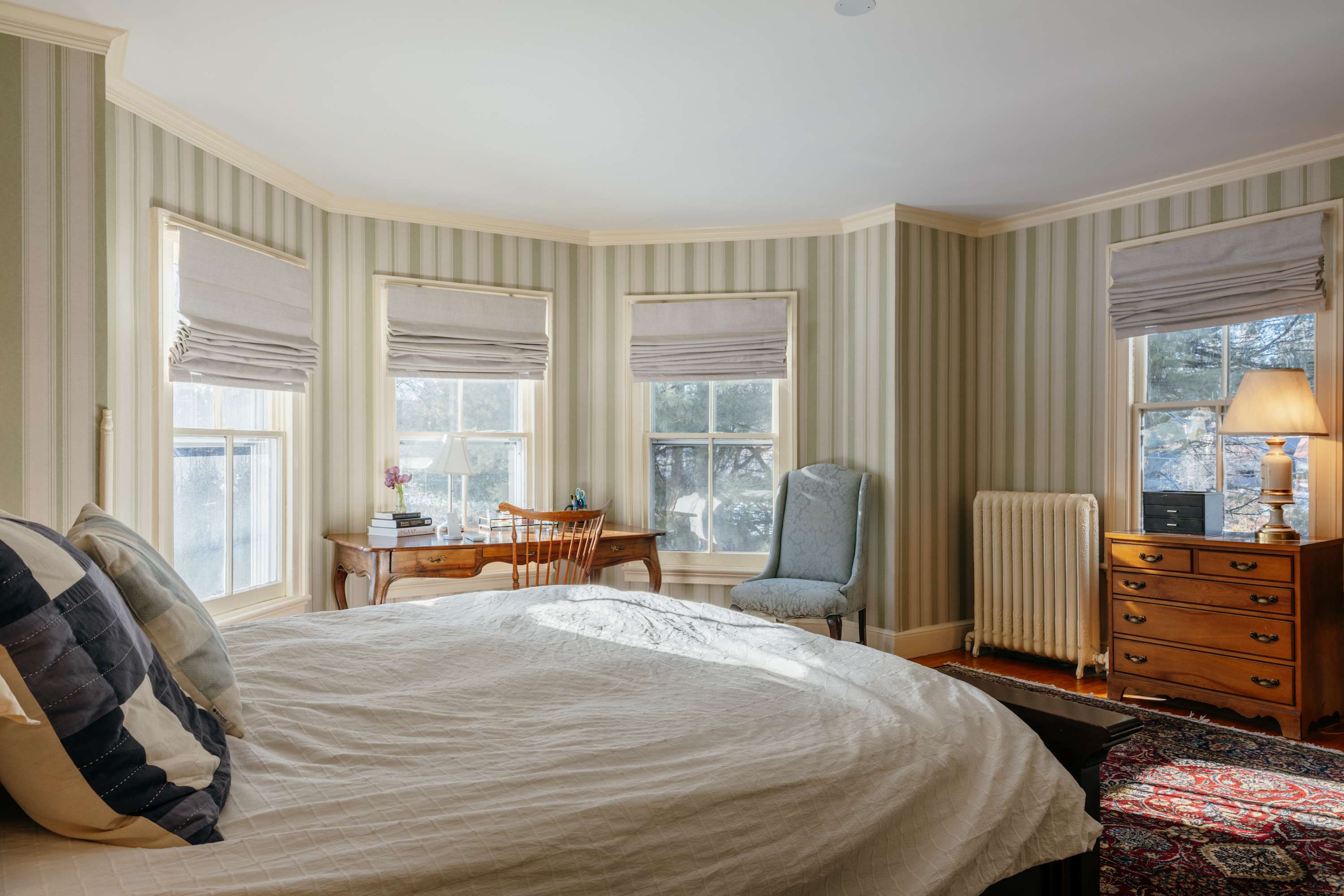 Spacious bedroom with light green and white striped wallpaper, a large bed with white bedding, and multiple windows fitted with light gray Roman blinds. A classic wooden desk and chair are positioned by the windows.