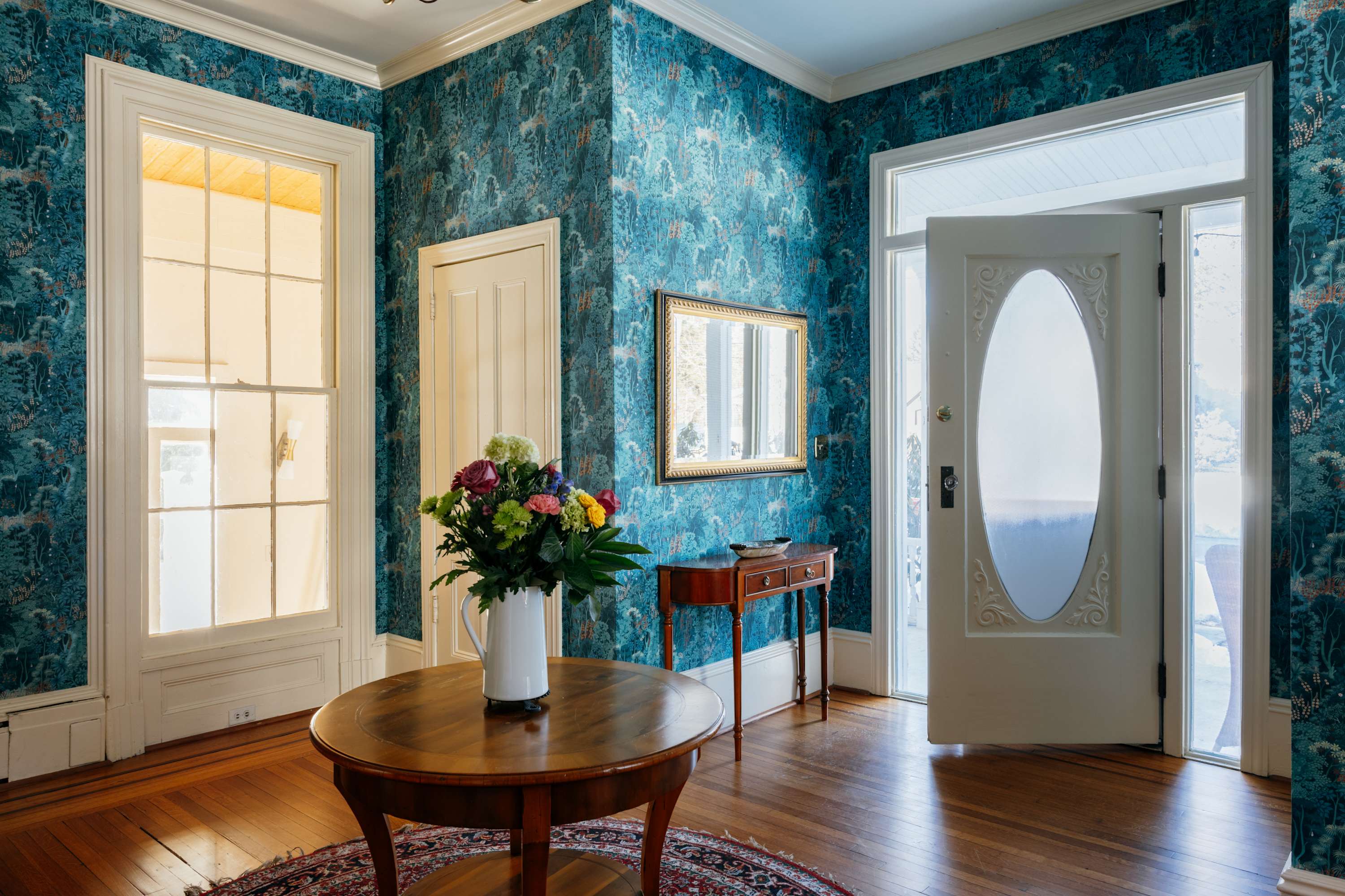 A grand foyer with vibrant blue patterned wallpaper, a round wooden table adorned with a fresh floral arrangement, and a large front door with an oval glass insert. Sunlight illuminates the polished hardwood floors and a tall window.