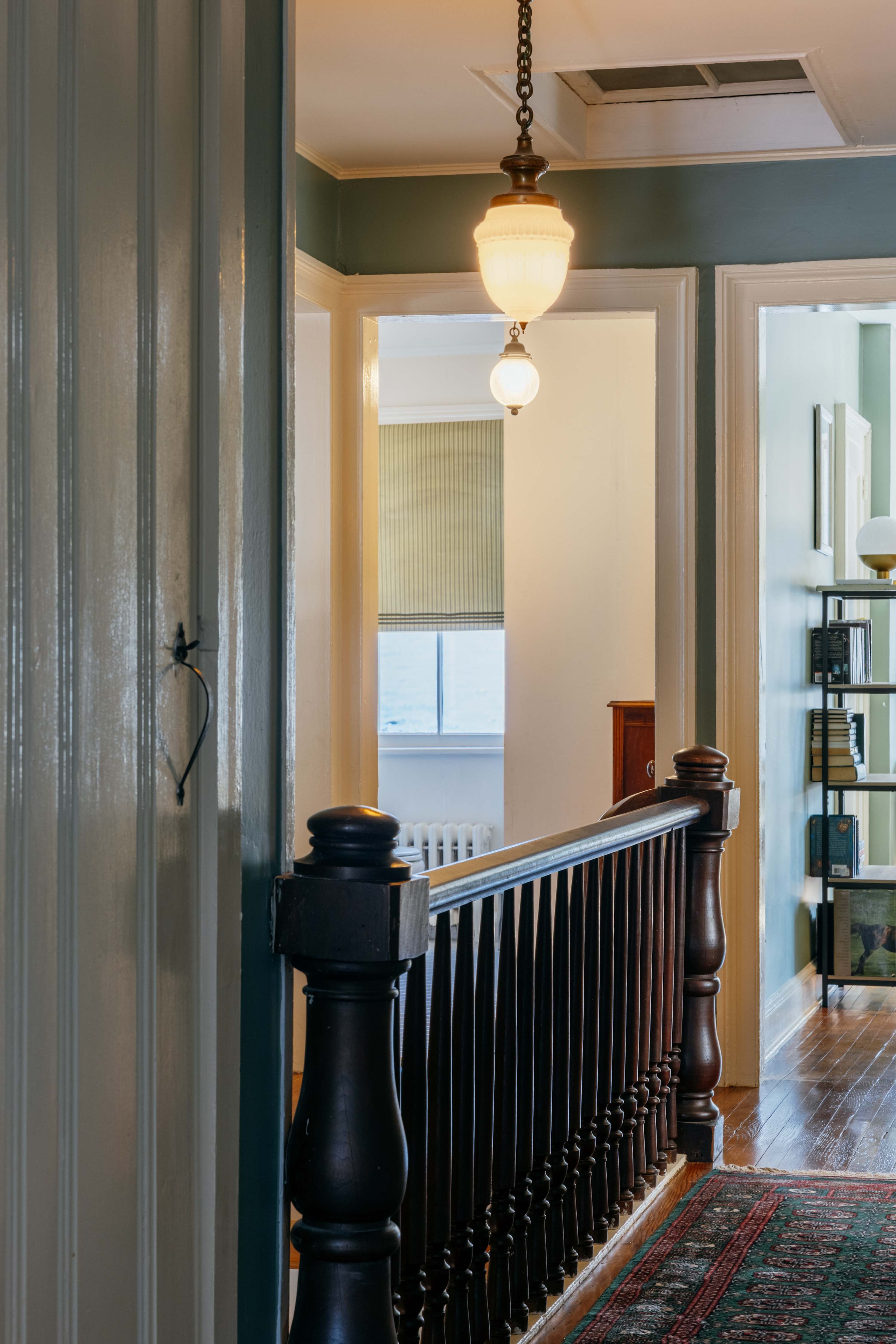 A classic staircase landing features a dark wooden banister and an ornate oriental rug. Two vintage pendant lights hang from the ceiling, illuminating doorways leading to other rooms with green walls.
