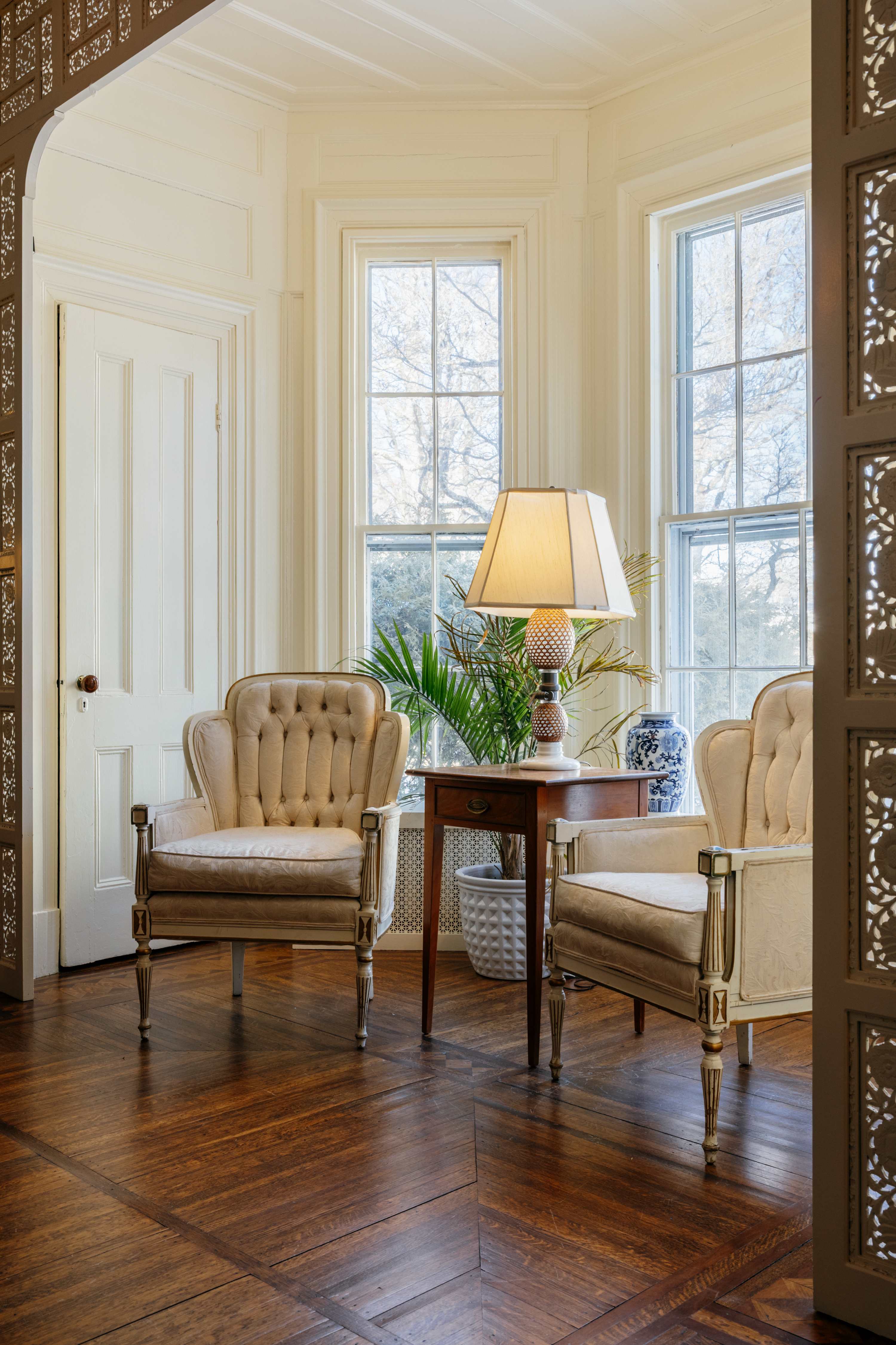 A bright sitting area with two elegant tufted armchairs flanking a small wooden side table with a lamp, set against large bay windows. The room features light-colored walls, intricate wooden floor patterns, and decorative carved screens.