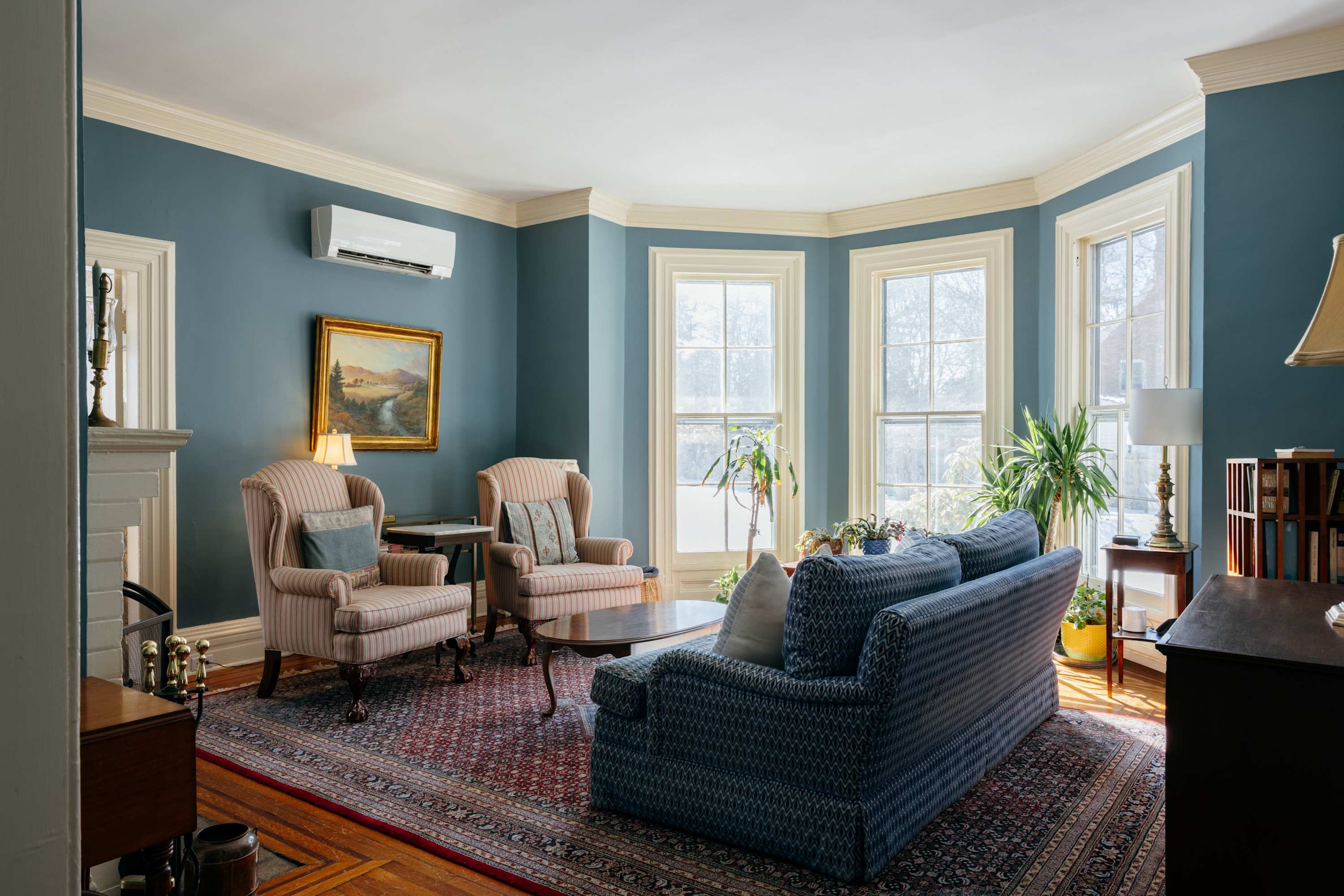 A spacious living room with deep blue walls, a patterned sofa, and two striped armchairs arranged around a coffee table. Large bay windows offer bright views, and the room is adorned with a colorful area rug and a painting above a fireplace.