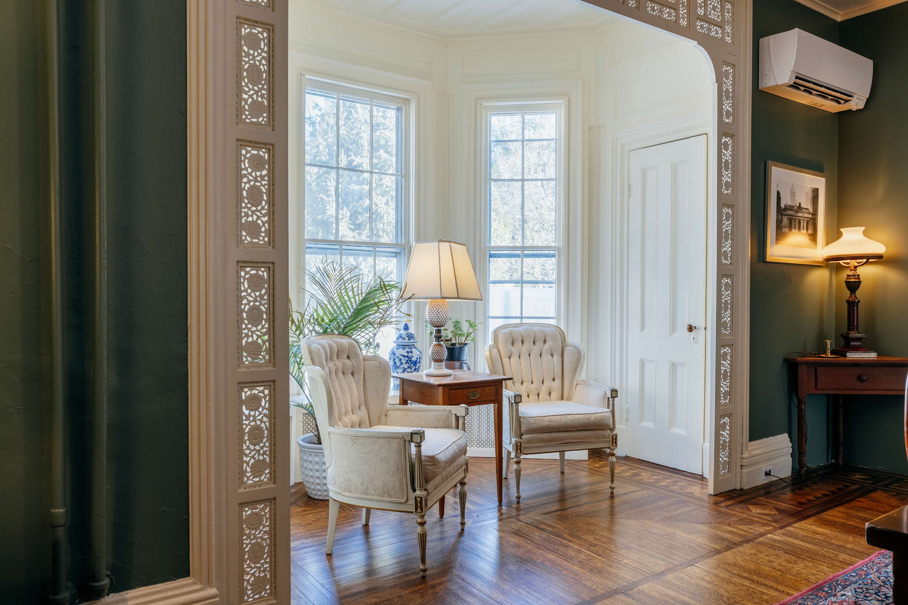 A bright sitting area with dark green walls and intricate white archways leading to a bay window with two upholstered armchairs and a small side table. Hardwood floors and a tall plant complete the elegant space.