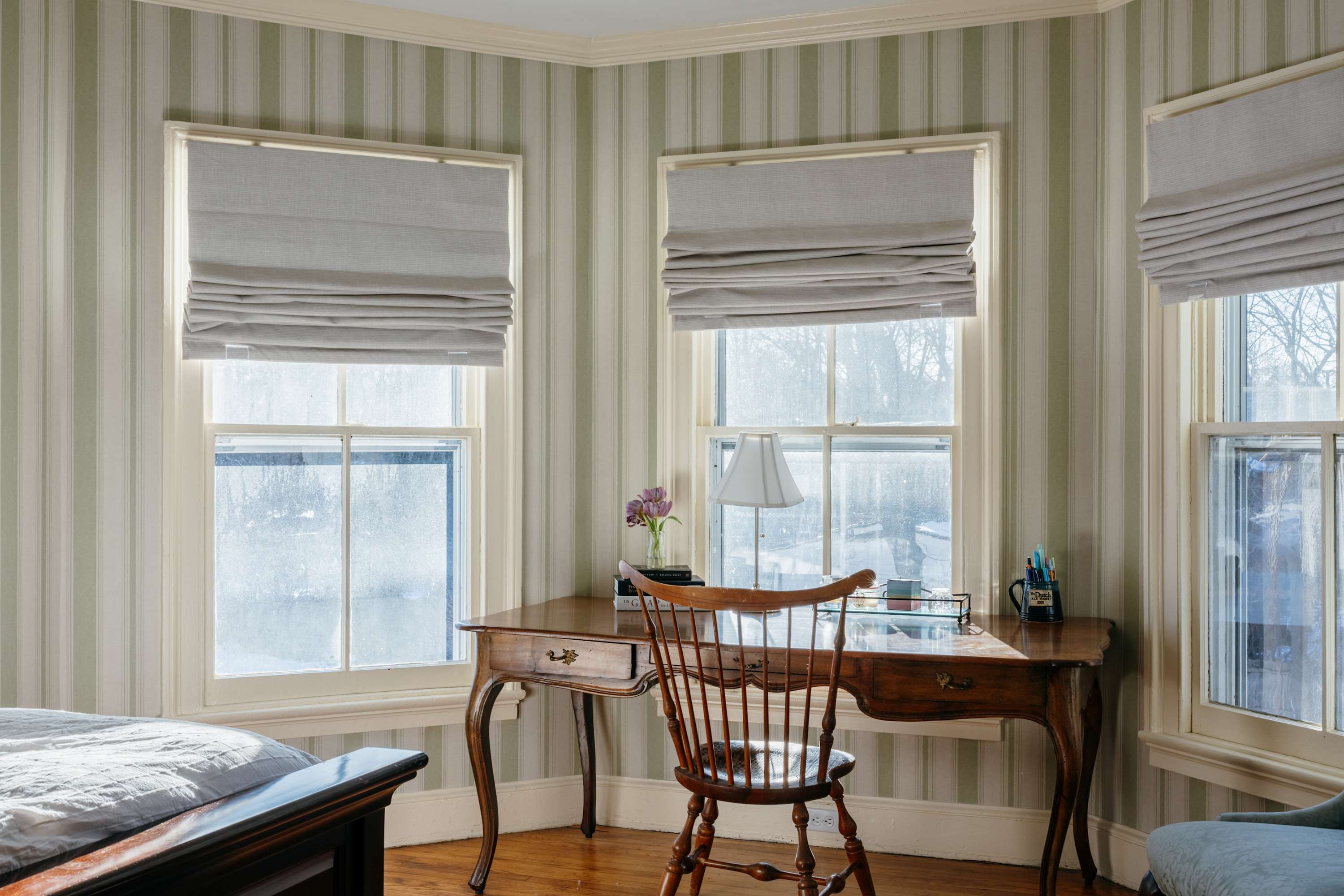 Detailed view of a wooden desk with a spindle-back chair in a bedroom with striped wallpaper. Multiple windows with light gray Roman blinds overlook a bright outdoor scene, with a table lamp on the desk.