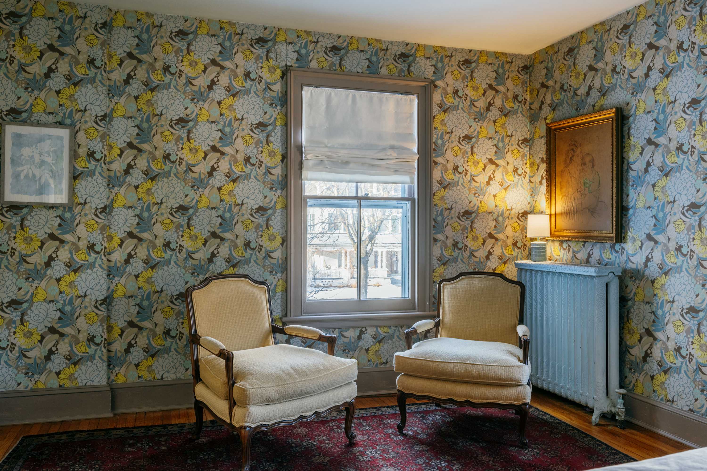 Cozy corner of a bedroom with vibrant floral wallpaper, featuring two upholstered armchairs positioned by a window with a white Roman blind. An ornate gold-framed painting hangs above a radiator.
