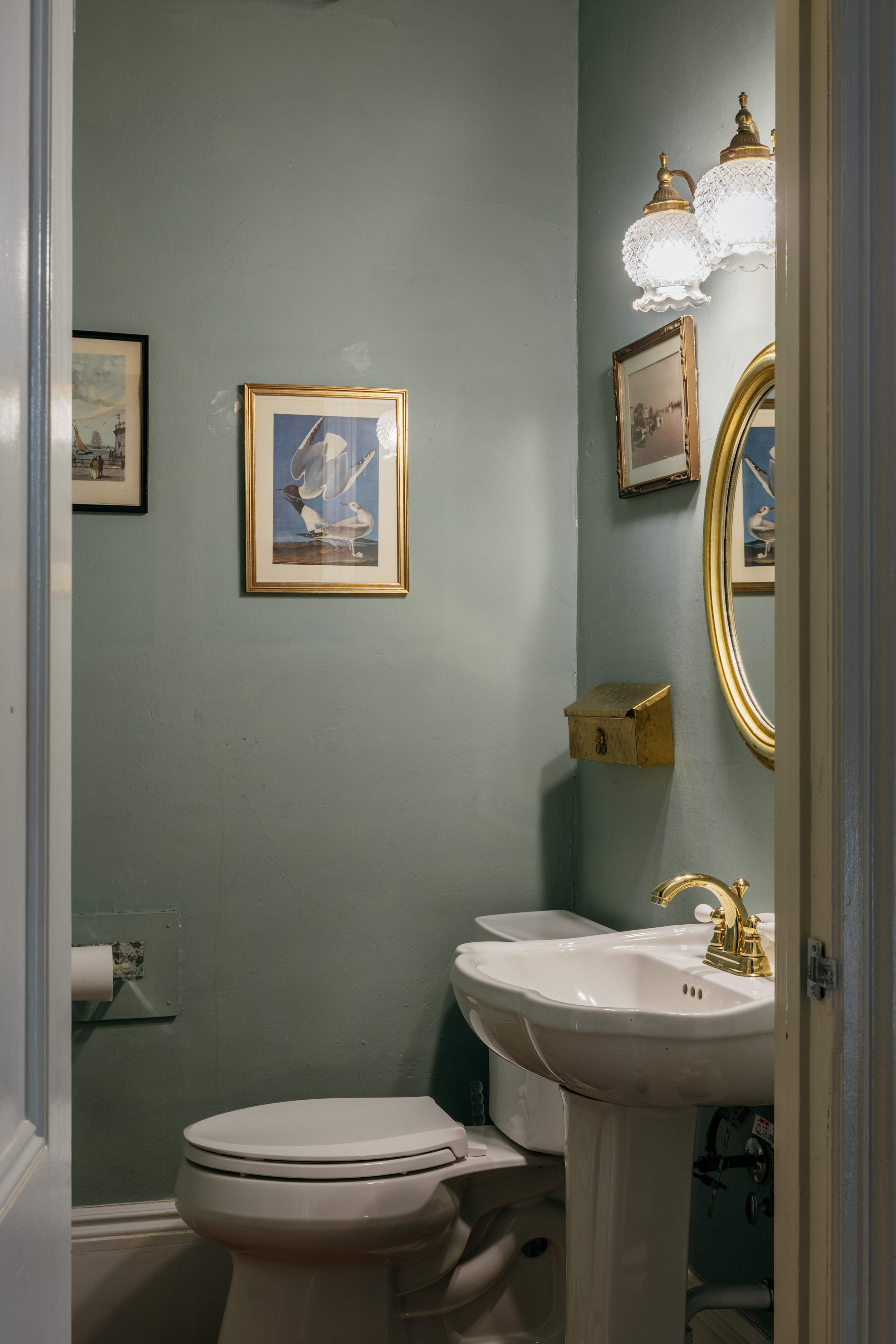 Compact powder room with a white pedestal sink, toilet, and a round gold-framed mirror. The walls are painted a muted blue-green, complemented by gold fixtures and ornate wall sconces.