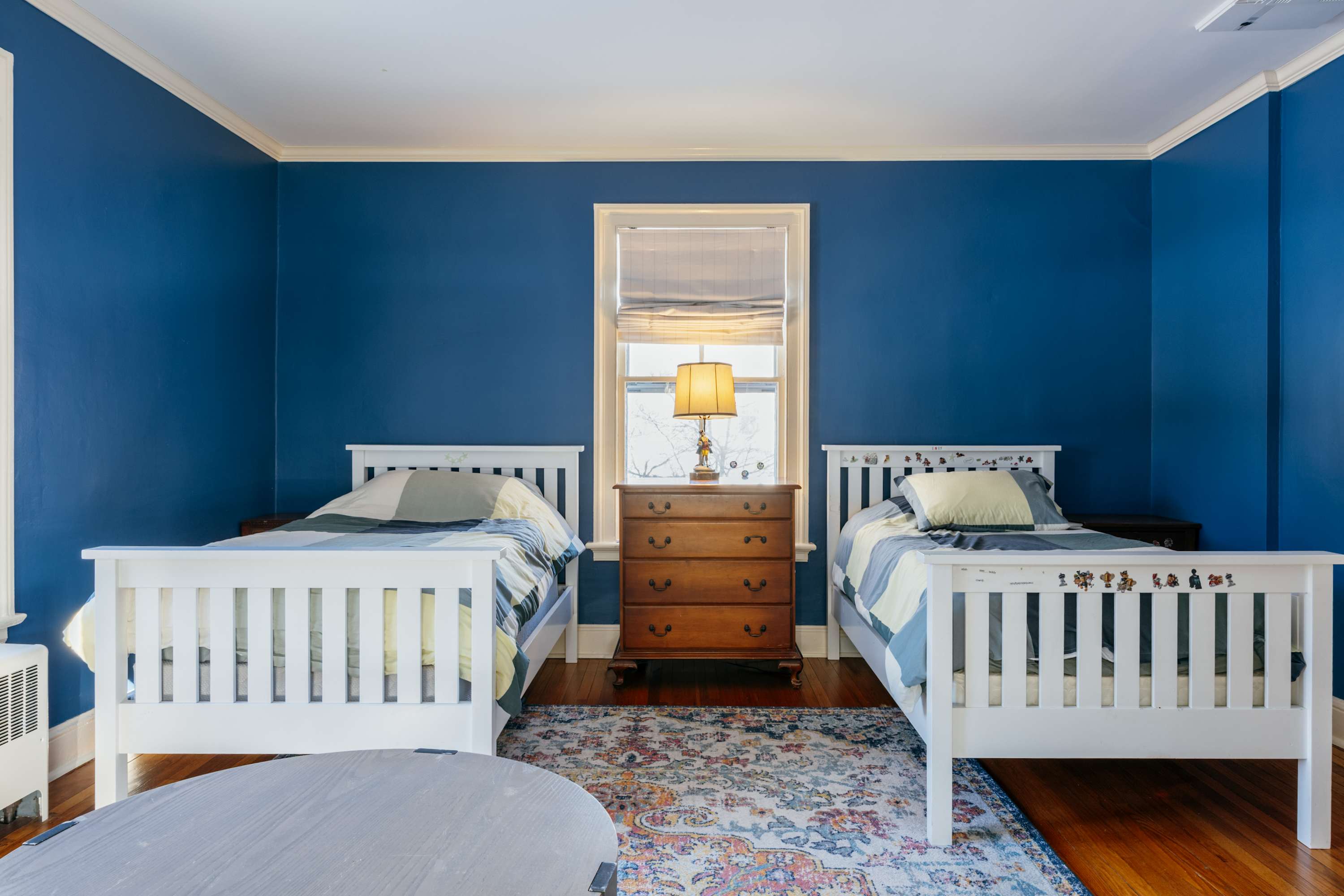 Vibrant bedroom with two white twin beds flanking a wooden dresser topped with a lamp. The room features bold blue walls, warm hardwood floors, and a large, ornate patterned area rug.