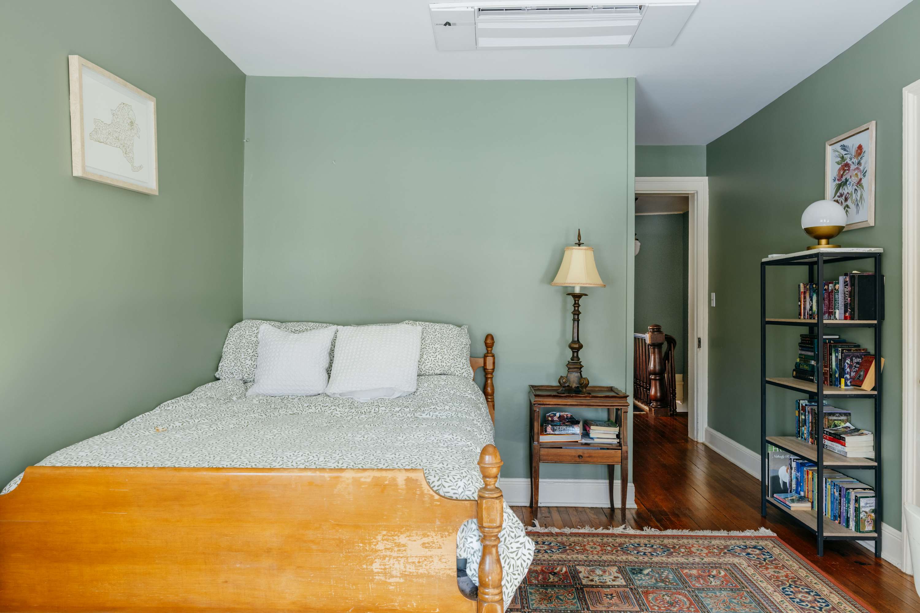 Cozy bedroom featuring a wooden bed with white bedding, a vintage nightstand, and a lamp. The room has light green walls, hardwood floors, and a colorful patterned area rug, with an AC unit visible on the ceiling.