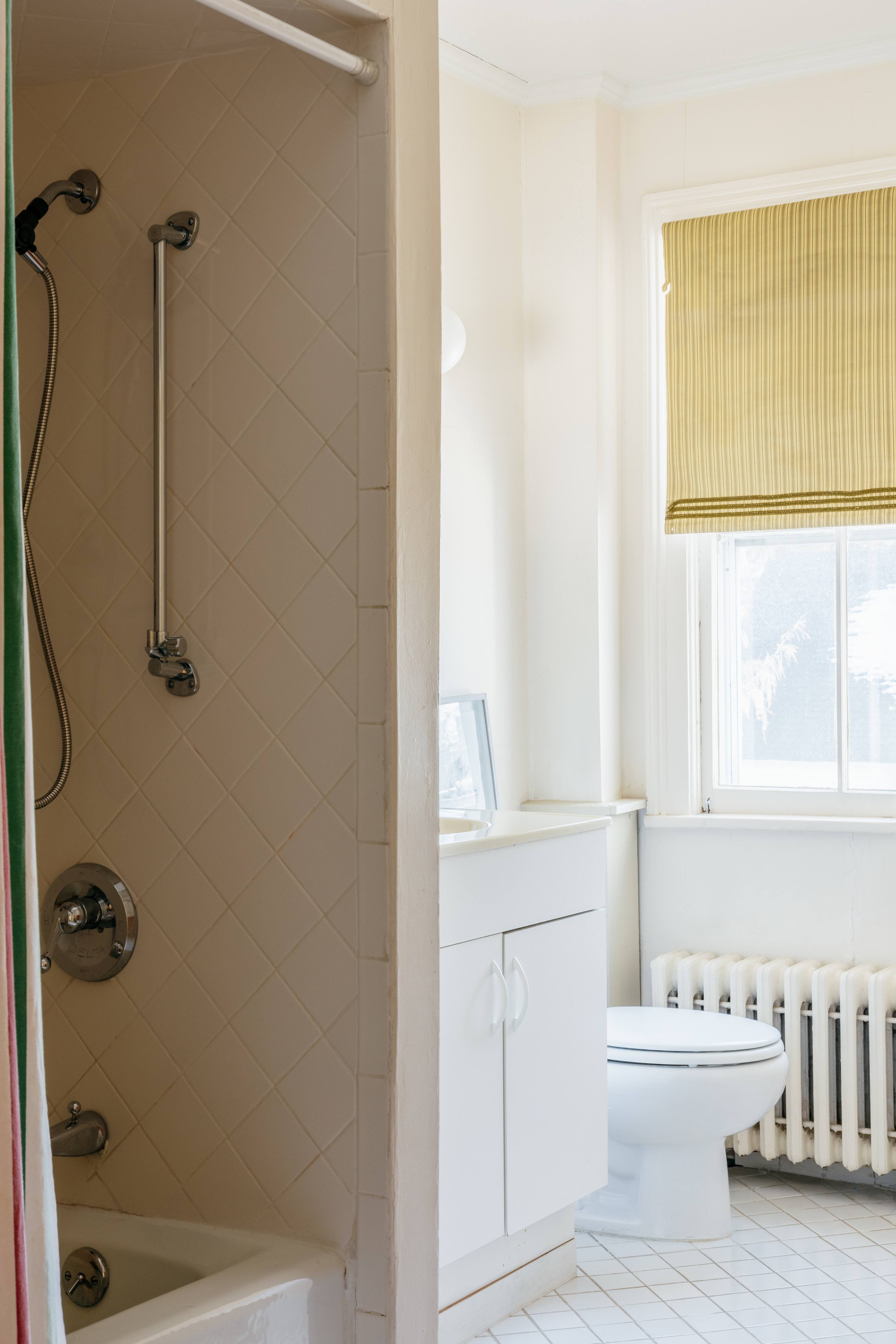 Bright bathroom with a shower-tub combination featuring white tiled walls and a colorful shower curtain. A white vanity and toilet are visible, with a window and yellow Roman shade above a radiator.