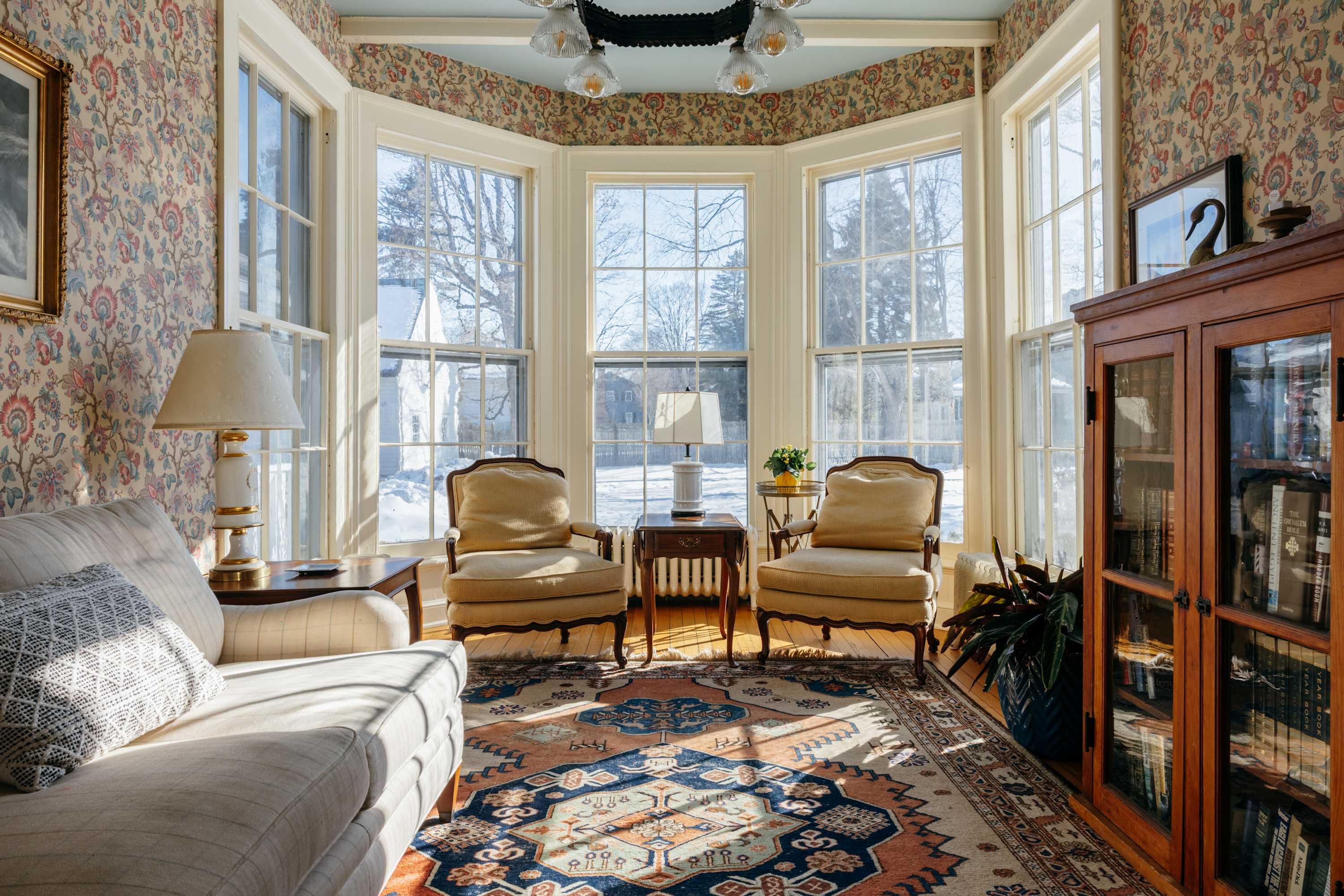 A sunlit living room with large bay windows offering views of a snowy landscape, floral wallpaper, and a patterned area rug. A white sofa and two yellow upholstered armchairs are arranged around a small wooden table.