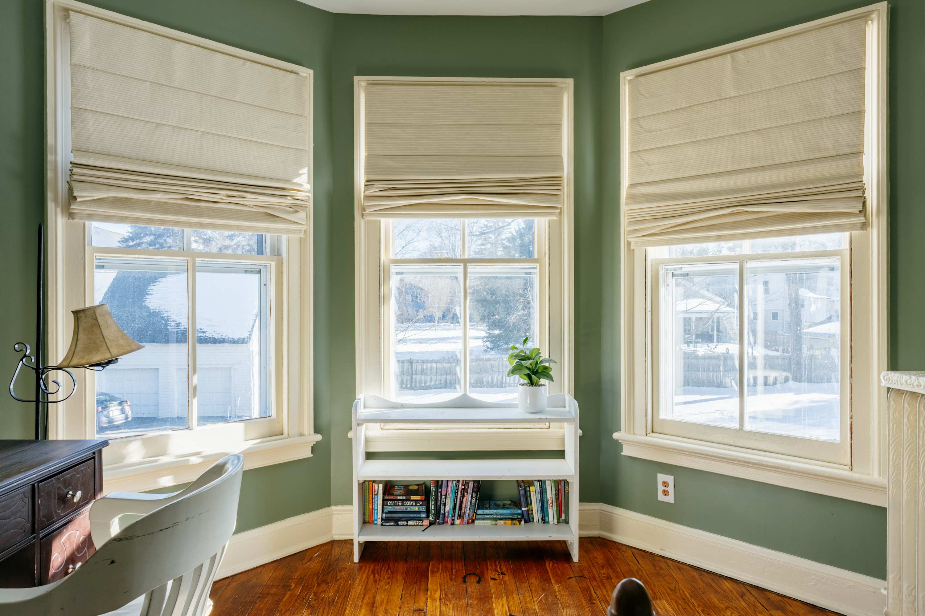 Bright bay window nook with three large windows, Roman shades, and a small white bookshelf filled with books. A desk and chair are partially visible on the left, set against sage green walls and warm hardwood floors.