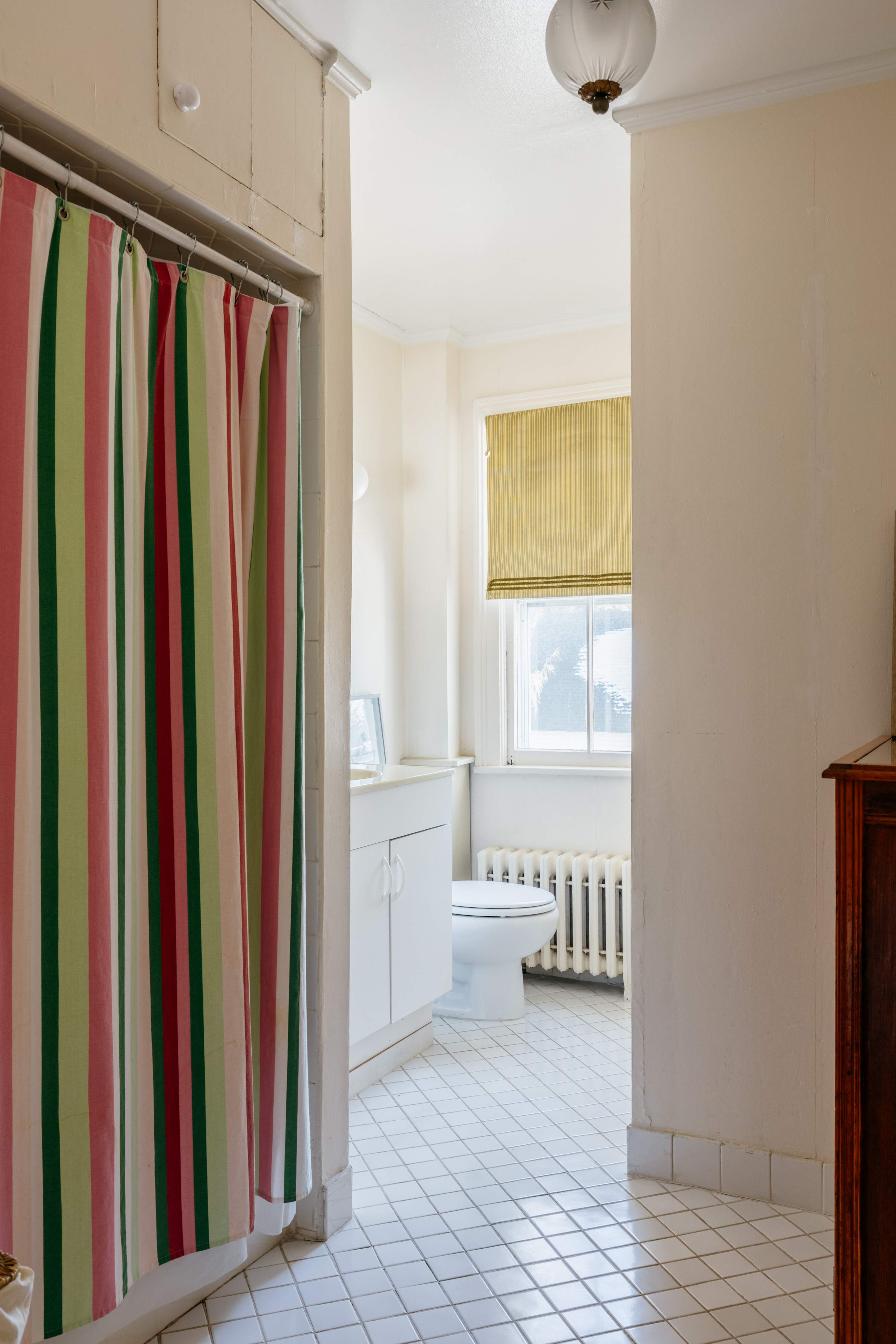 View of a bathroom featuring a vibrant striped shower curtain enclosing a shower-tub combo. A white vanity, toilet, and window with a yellow Roman shade are visible, set against white tiled floors.