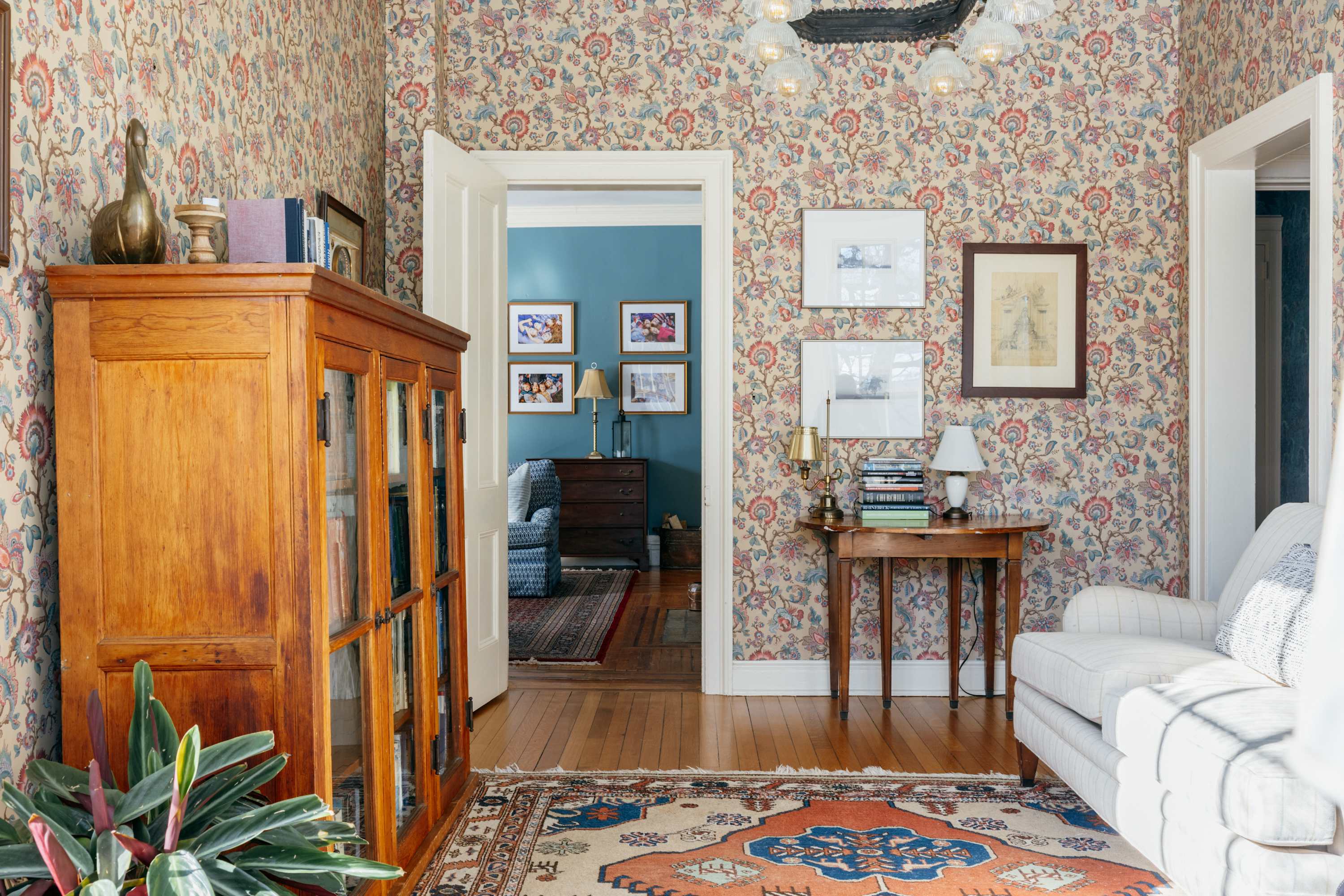 A cozy living space with floral wallpaper and hardwood floors, featuring a white sofa and a wooden display cabinet. An open doorway leads to another room with blue walls, suggesting a connected layout.