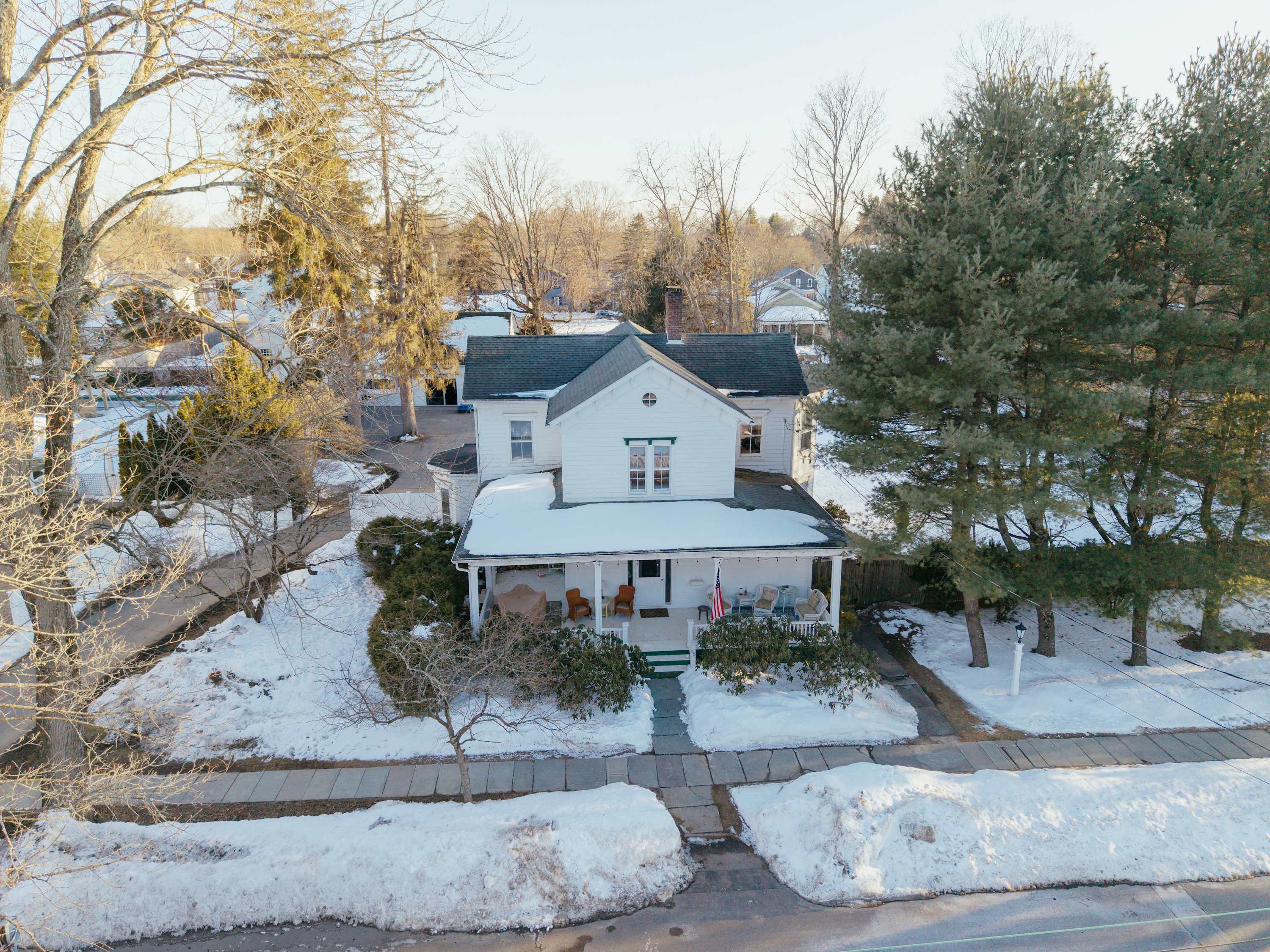 Aerial view of the white house in winter, featuring a snow-covered front yard and a welcoming covered porch. Bare trees surround the property under a bright sky.