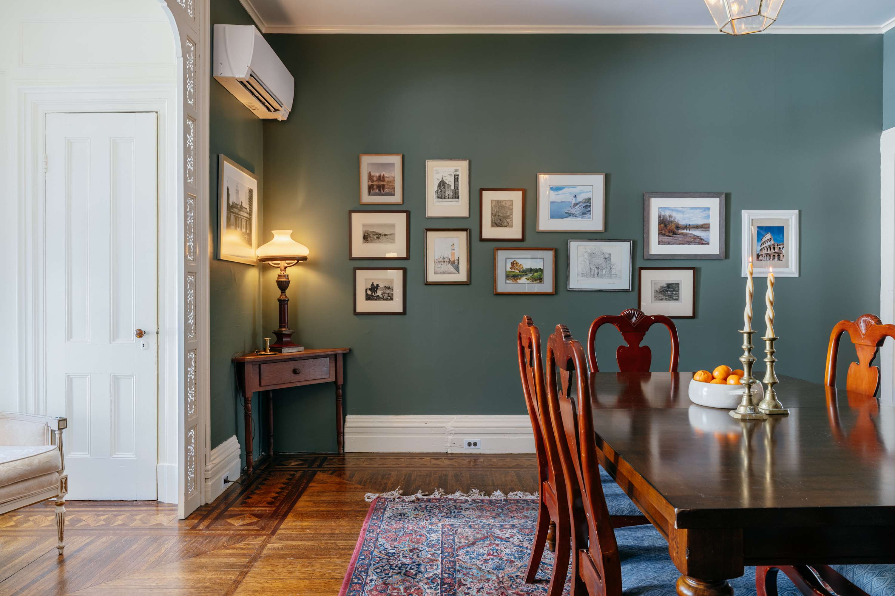 An elegant dining room featuring dark green walls adorned with a gallery wall of framed artwork, a long polished wooden dining table with classic chairs, and a decorative area rug. A mini-split AC unit is visible above a side table with a lamp.
