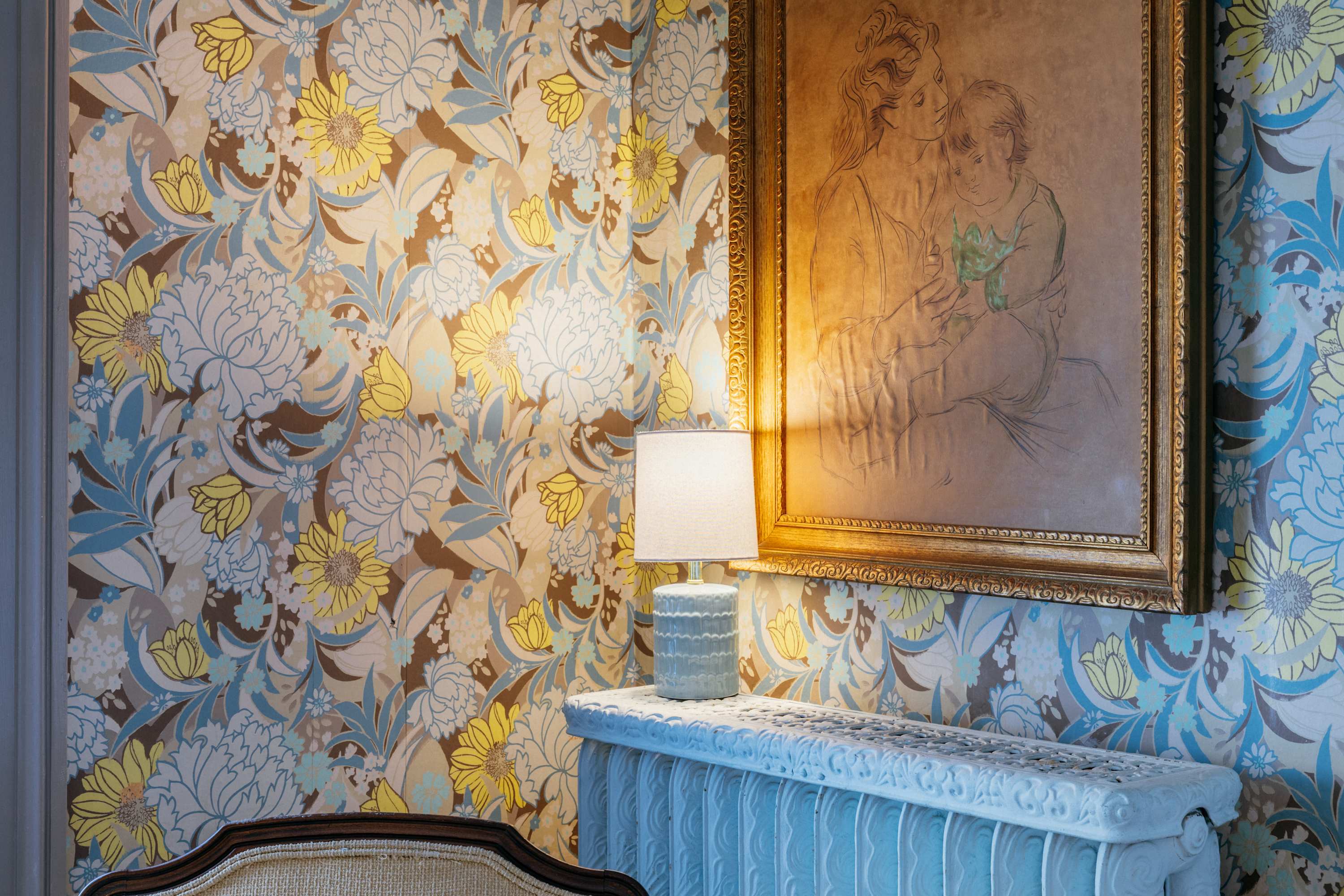 Eclectic living space featuring bold floral wallpaper in yellow, blue, and brown tones. A large framed artwork hangs above a decorative white radiator cover, illuminated by a small table lamp.