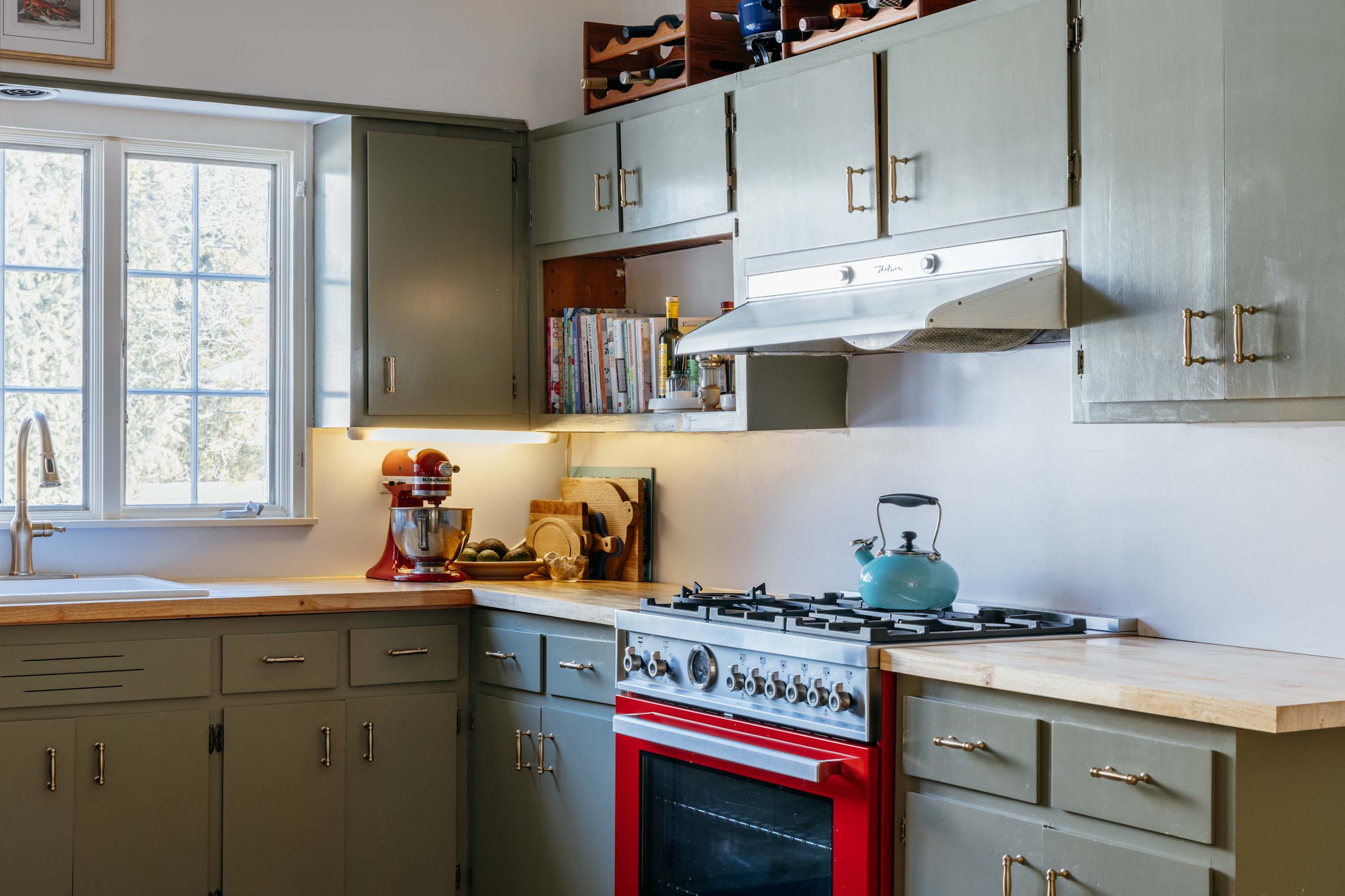 A functional kitchen area with sage green cabinets, light wood countertops, and a prominent red gas range with a stainless steel hood. Open shelving displays cookbooks, and a window provides natural light over the sink area.