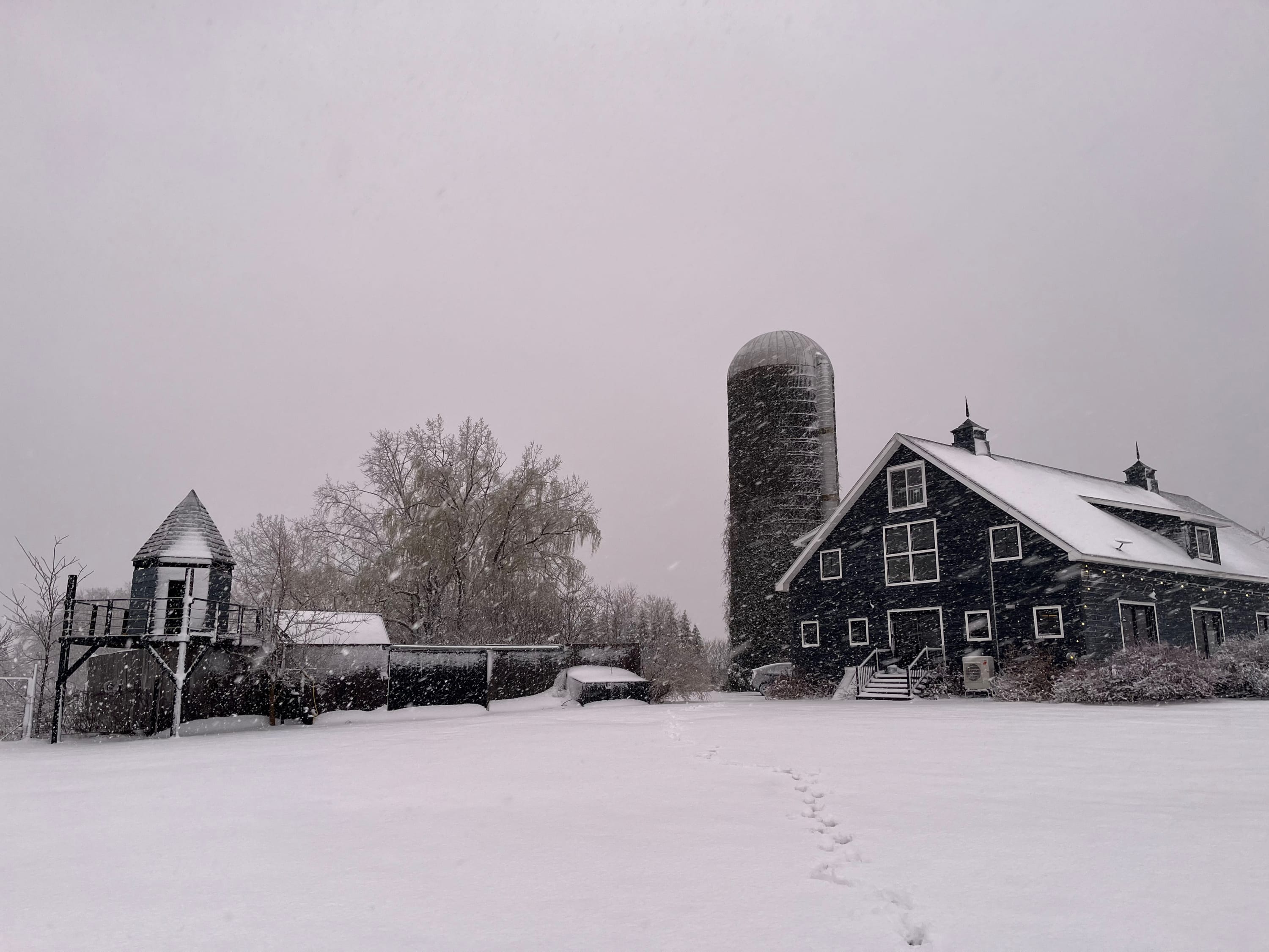 A wide shot of the dark gray barn-style house and a tall silo, both covered in snow, with a whimsical treehouse structure to the left, all under a soft, snowy sky.