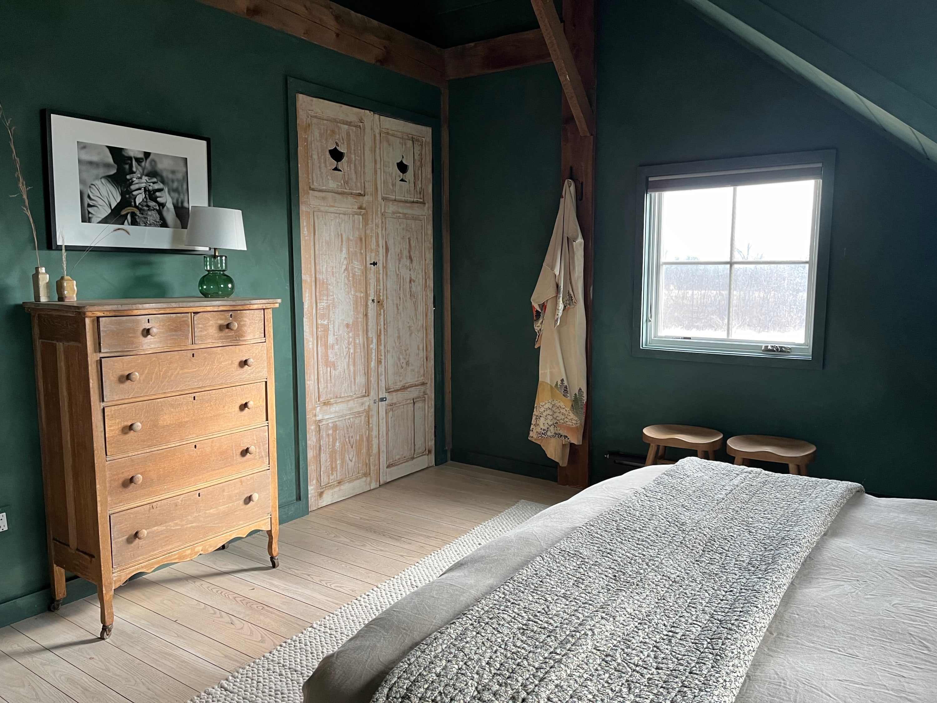 A close-up view of the green-walled bedroom, showcasing a vintage wooden dresser with a lamp and framed art, a rustic wooden closet, and a bed with a textured throw blanket.