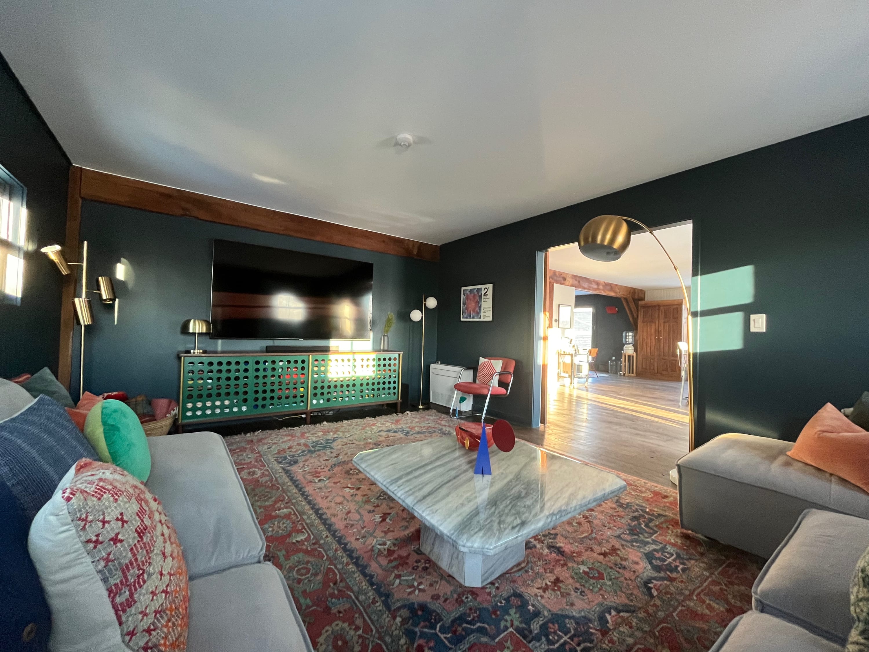 Modern living room with dark green walls, a large flat-screen TV mounted above a vibrant green console, a light gray sectional sofa, and a large patterned rug, with sunlight streaming in from windows.