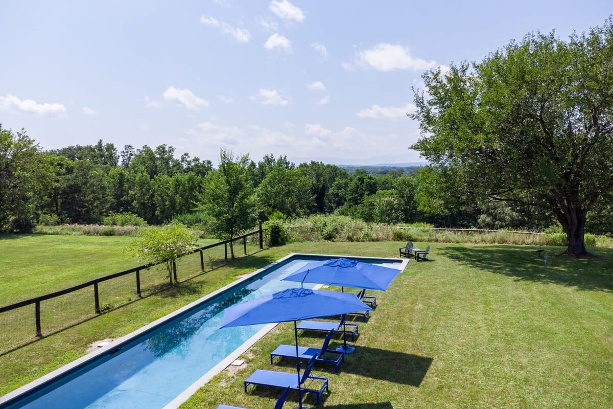 An aerial view of a long, narrow swimming pool with blue lounge chairs and umbrellas on a vibrant green lawn, surrounded by a black fence and dense green trees in the distance under a bright blue sky.