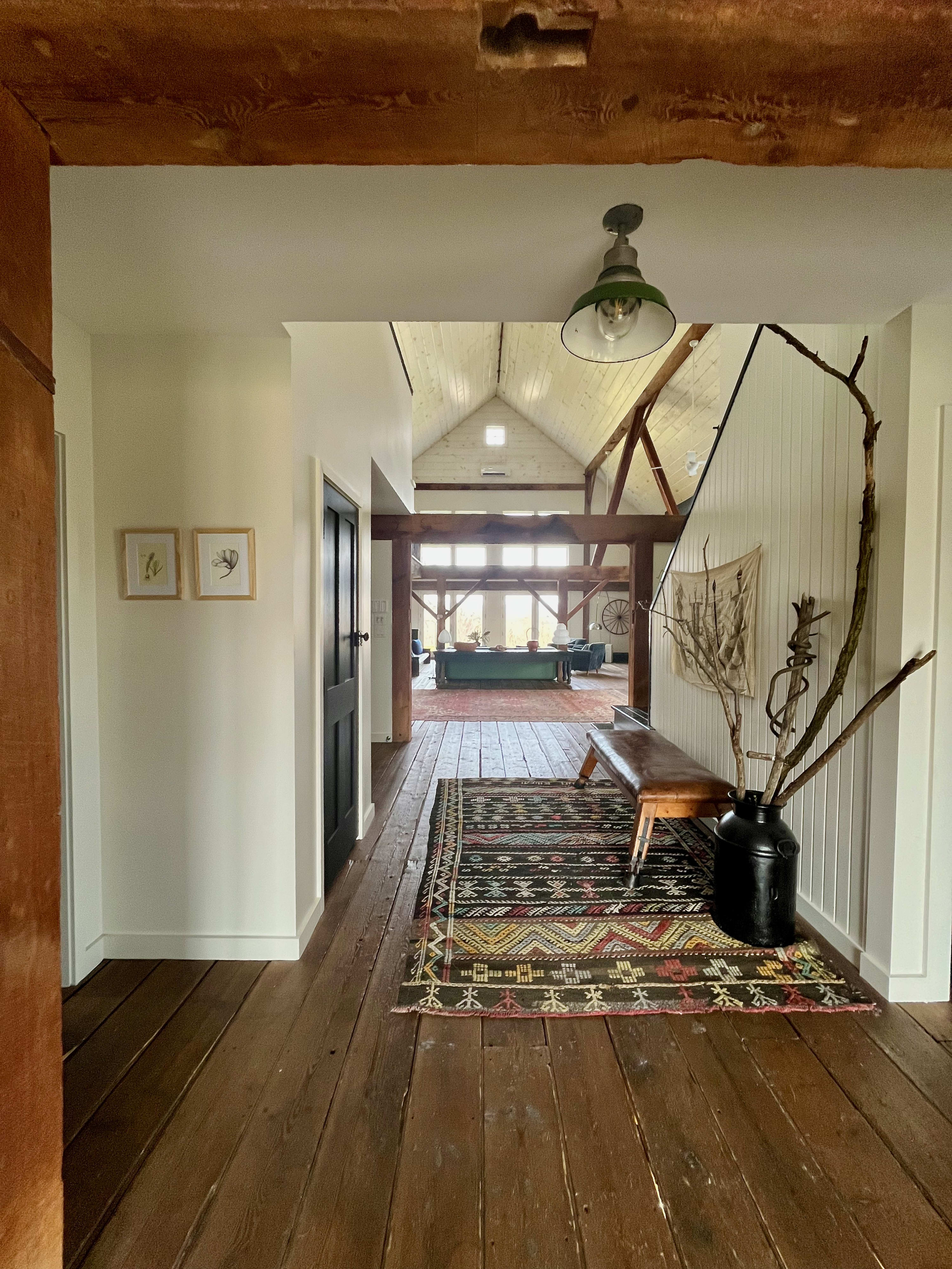 A rustic hallway with wide-plank wood floors leads towards a vaulted ceiling living area with exposed beams. A wooden bench, a large black vase with branches, and a colorful patterned rug decorate the space.