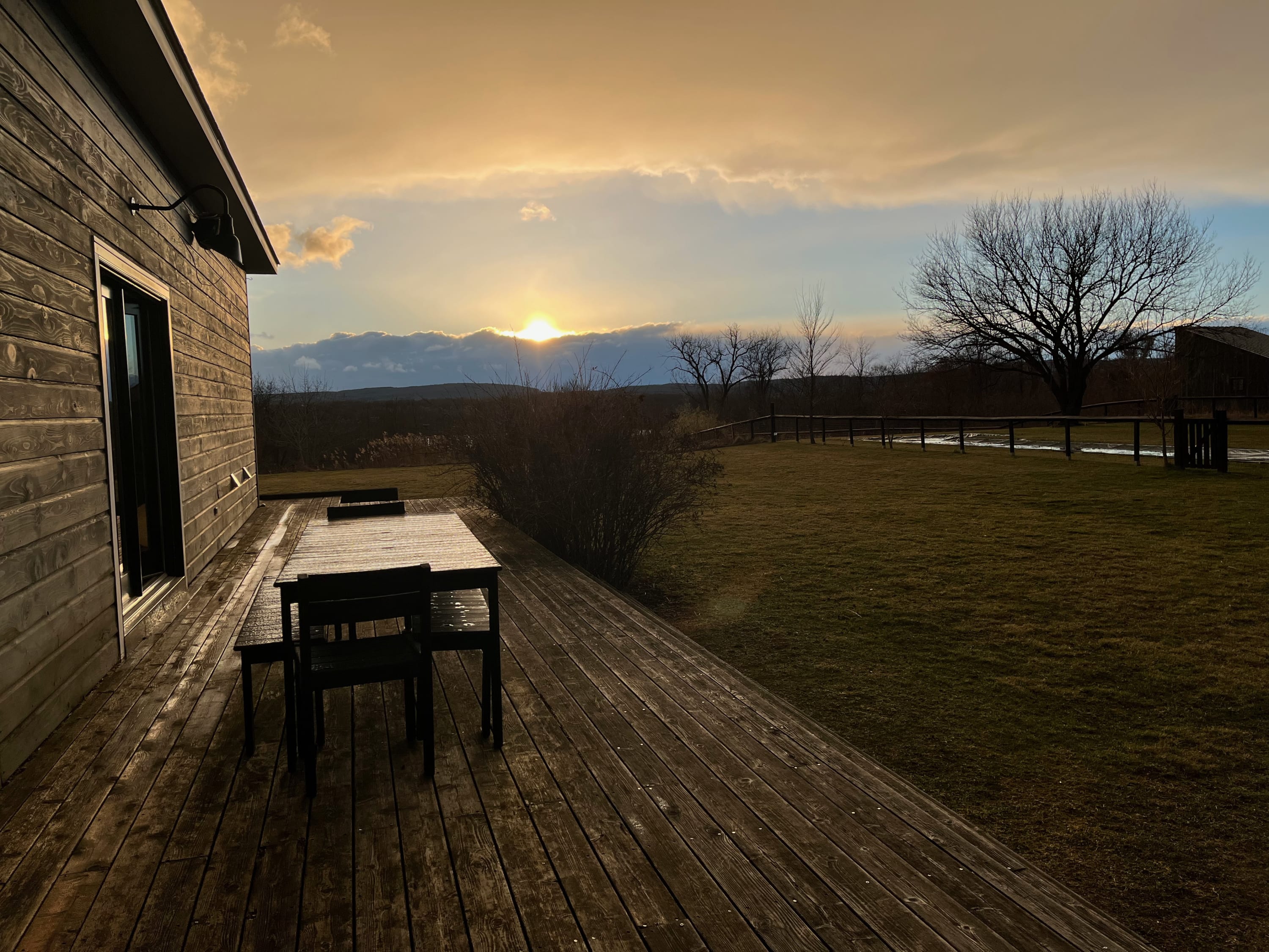 Outdoor wooden deck with a picnic table and benches, overlooking a grassy yard and a scenic landscape with a sunset, surrounded by natural vegetation.