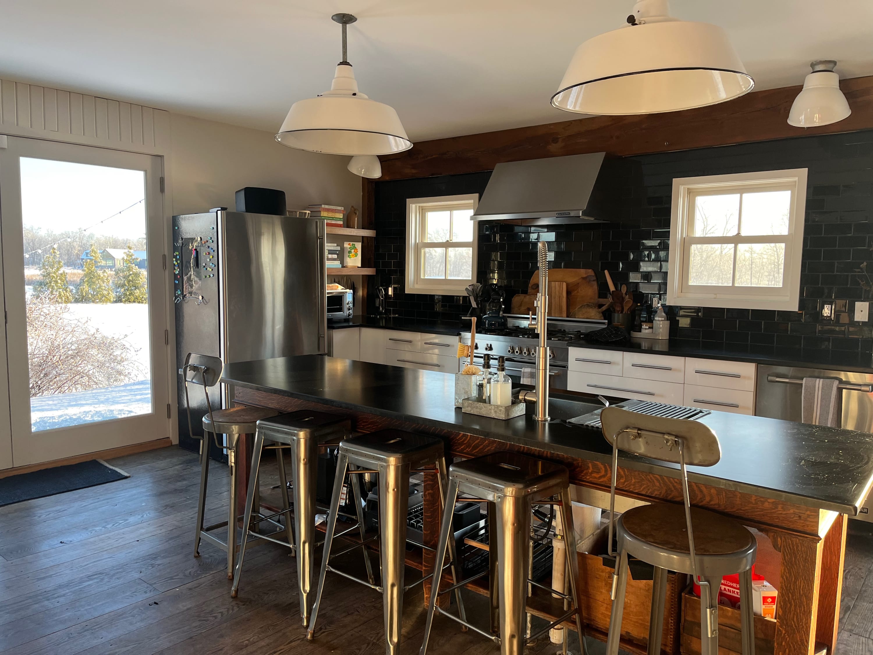 Another bright view of the kitchen, showcasing the large stainless steel refrigerator, a wooden island with a dark countertop and metal stools, and light wood floors. Exposed wooden beams and white pendant lights add to the rustic-modern aesthetic.