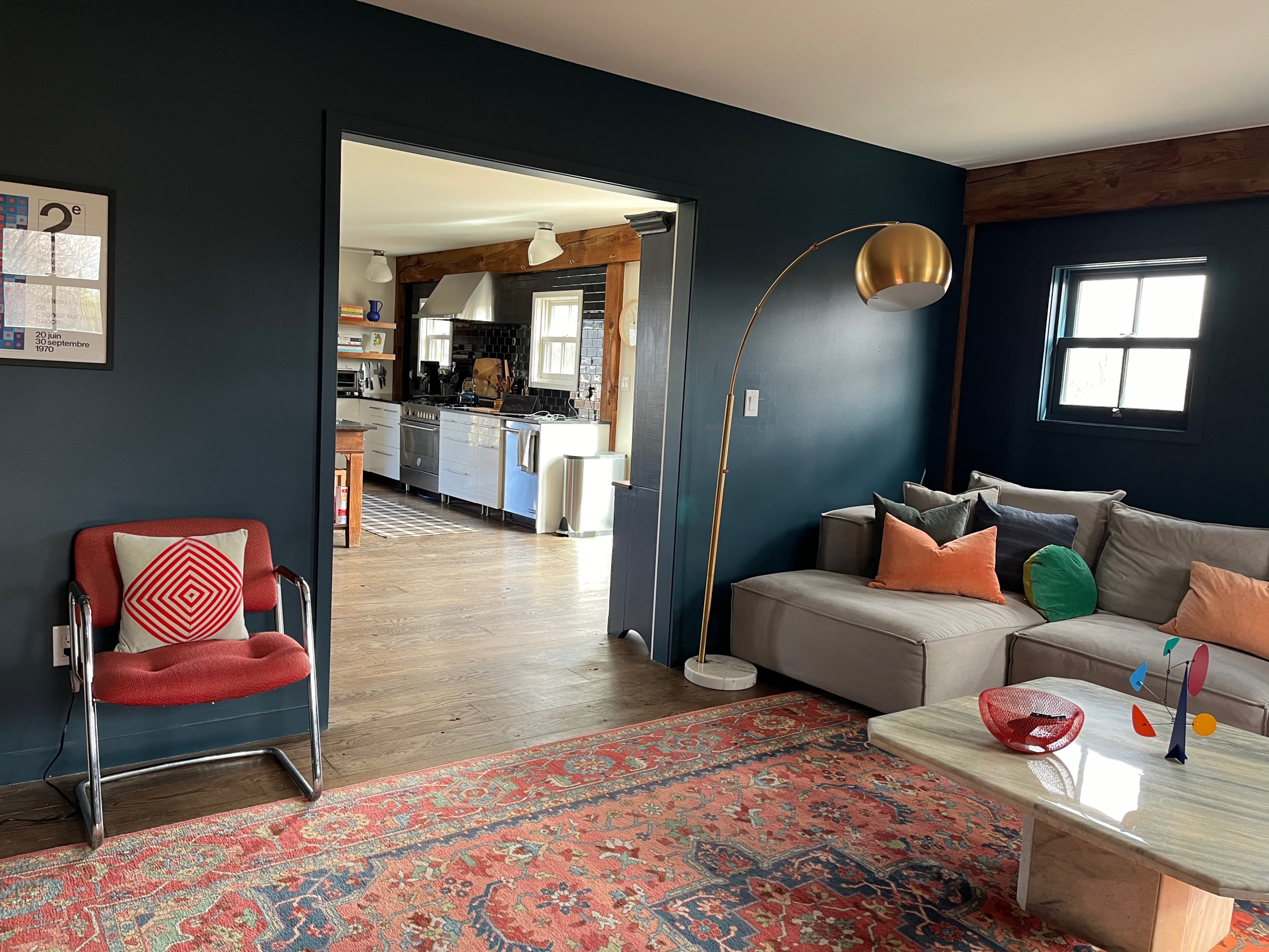 A cozy living room with dark blue walls, a light gray sectional sofa, and a patterned red and blue area rug, offering a glimpse into a modern kitchen through an open doorway.