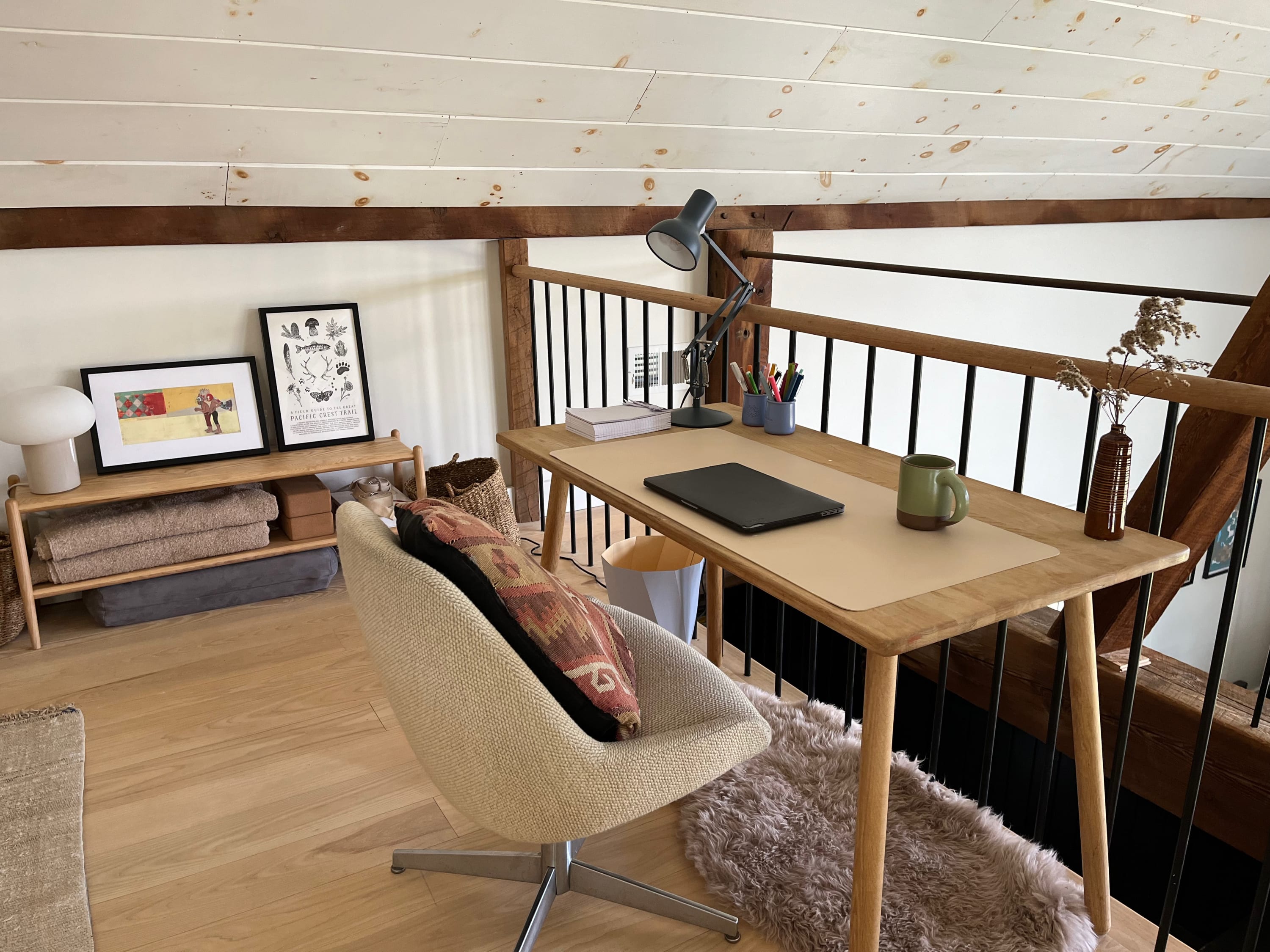 Close-up of the loft's office nook, showcasing a light wood desk with a task lamp and an ergonomic chair with a patterned pillow. A small wooden shelf with decor and a fluffy rug complete the inviting workspace.