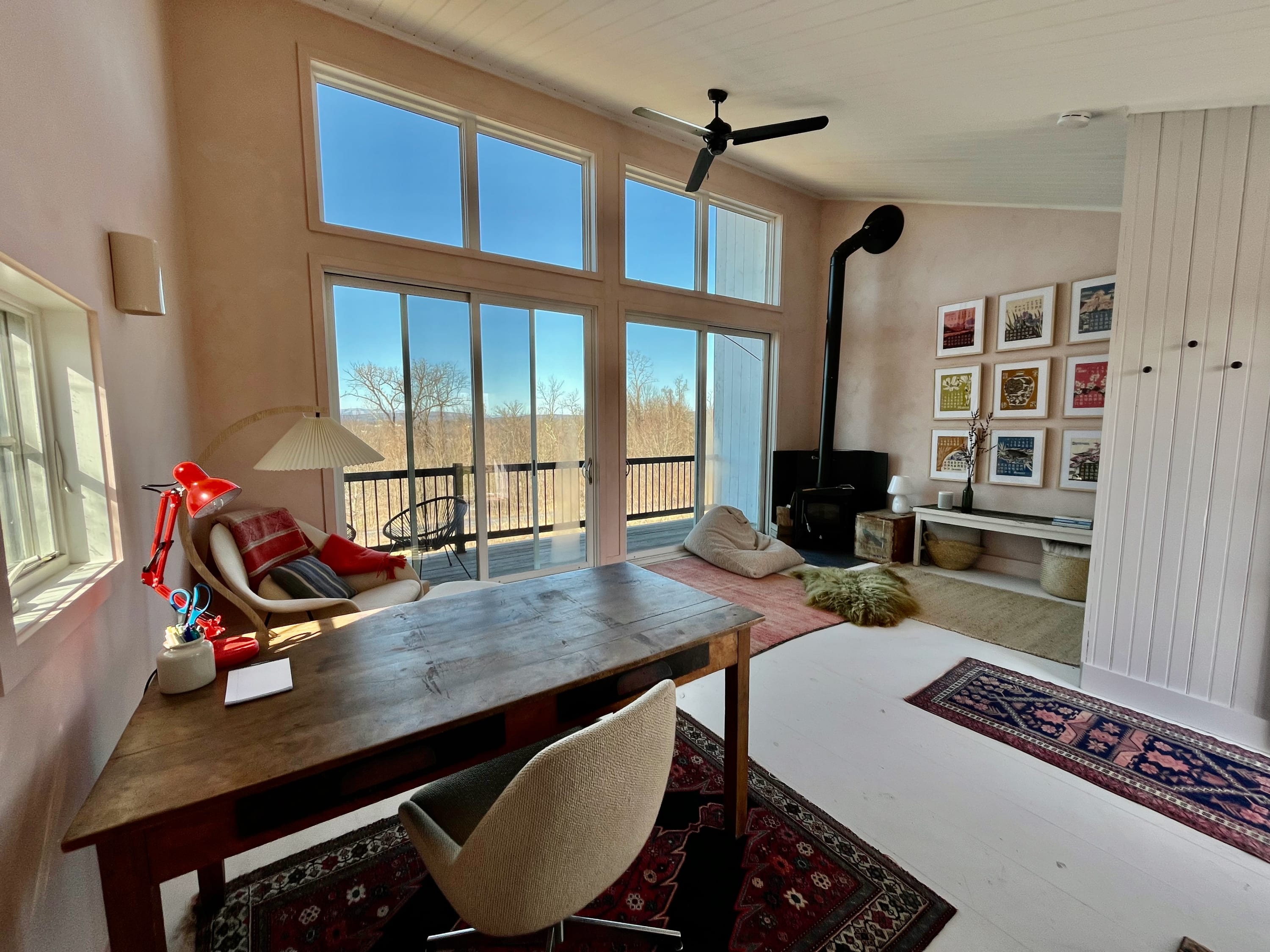 Expansive living space with light pink walls, a high white planked ceiling, and large windows offering panoramic views, featuring a wood-burning stove, a wooden desk, and a ceiling fan.
