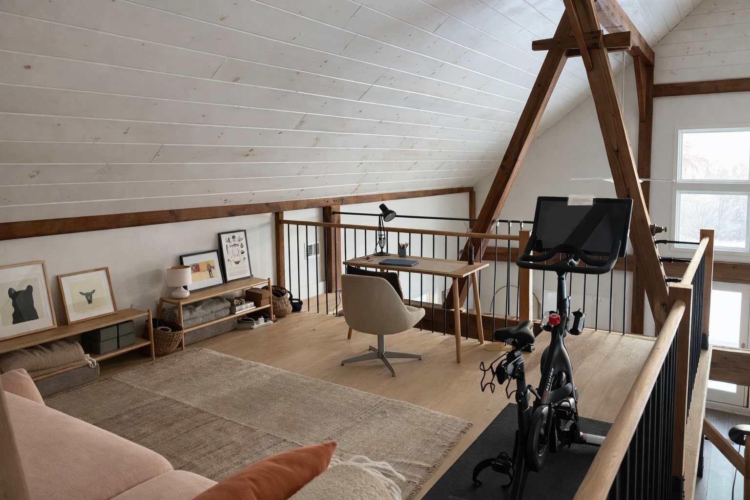 Spacious loft area with a dedicated office space featuring a wooden desk, ergonomic chair, and a Peloton bike. Exposed wooden beams and a slatted white ceiling add architectural interest, overlooking a lower level.
