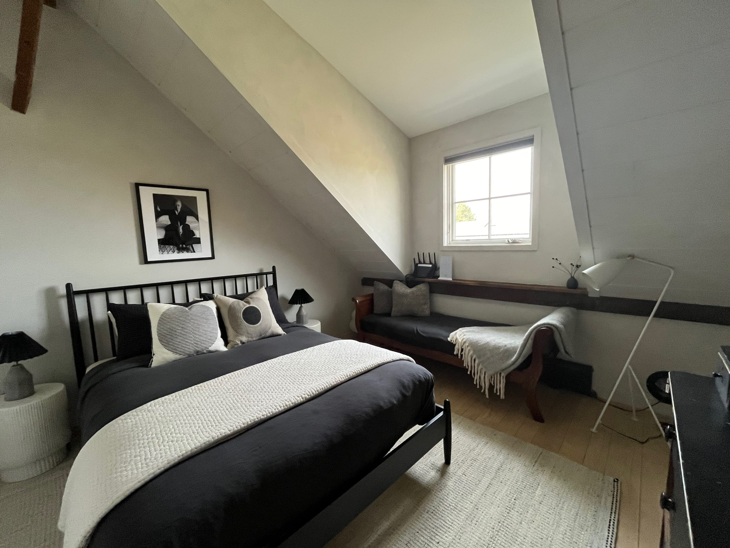 Cozy bedroom with a black metal bed frame, dark bedding, and a white textured rug. Slanted ceilings and a small window give it an attic-like feel, with a daybed and a small side table visible.