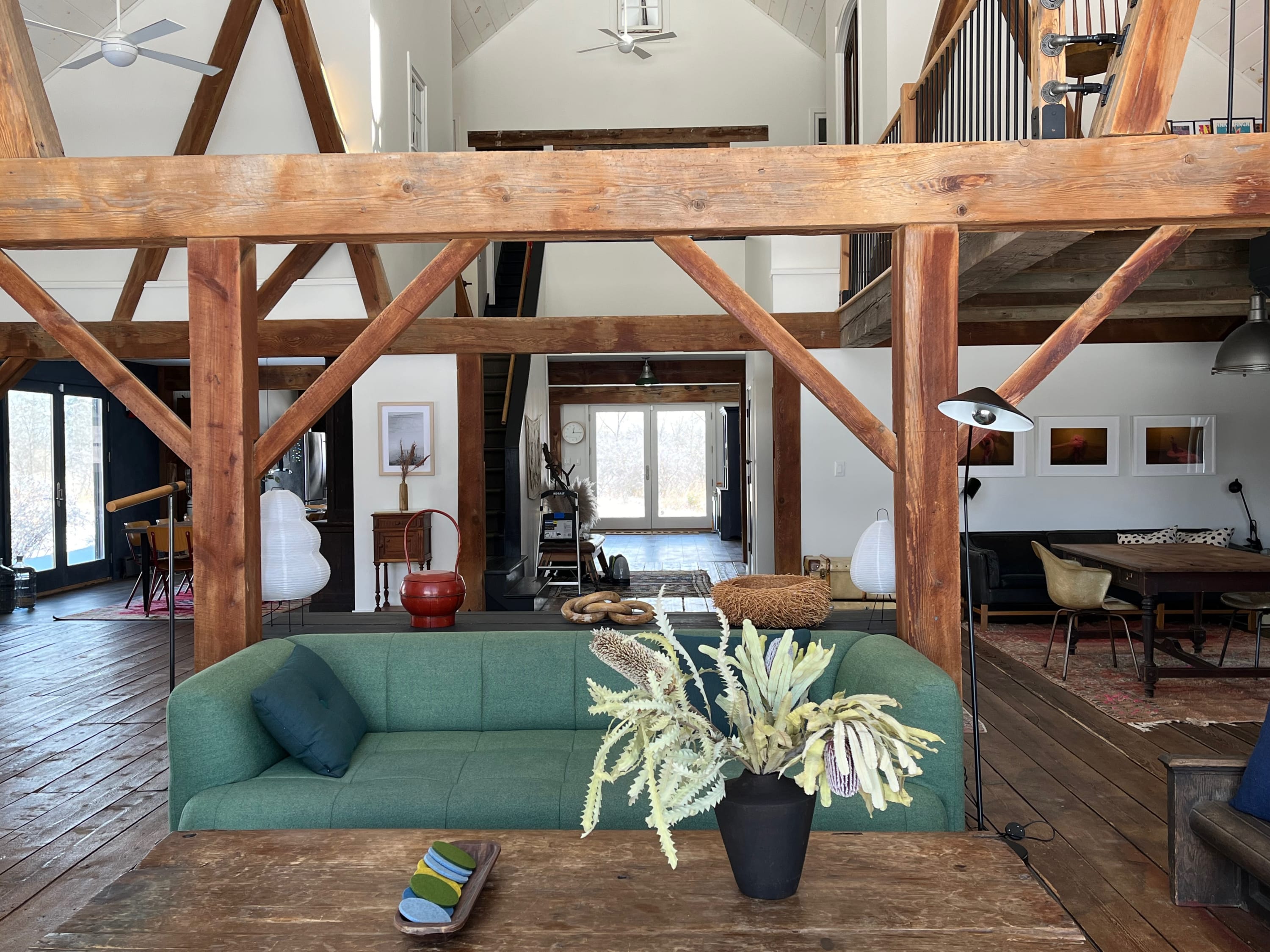 A spacious living area with exposed wooden beams and hardwood floors features a green sofa, a rustic wooden coffee table adorned with dried floral arrangements, and a view into other parts of the open-concept barn conversion.