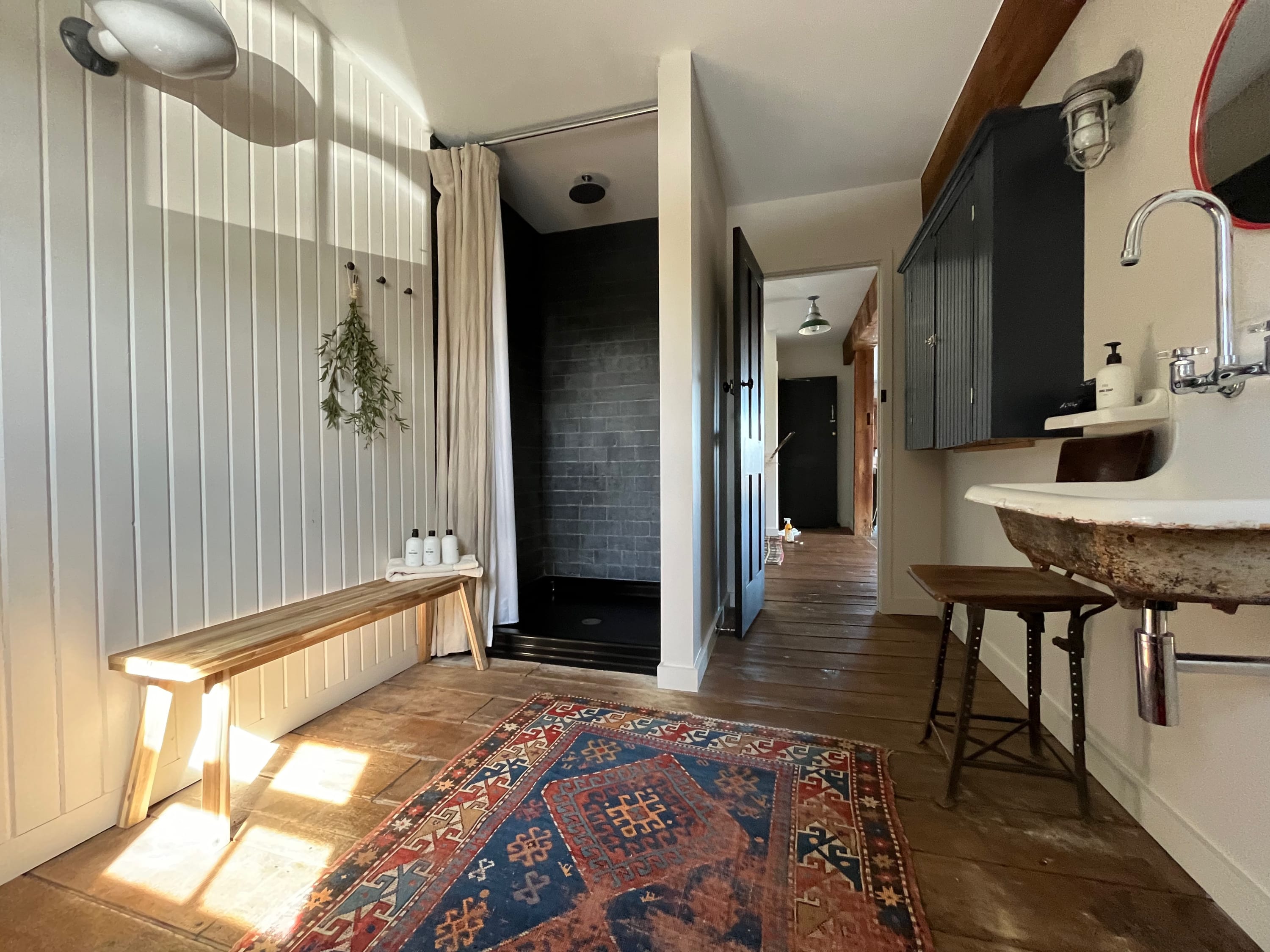 This rustic bathroom features a walk-in shower with black subway tiles and a light curtain, a wall-mounted sink, and a wooden bench. Wide-plank wood floors and white vertical paneling create a bright, clean aesthetic.