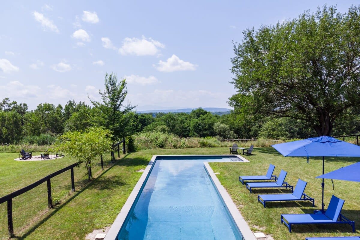 Elevated view of a long, narrow blue swimming pool with blue lounge chairs and umbrellas on a green lawn, overlooking a vast expanse of green trees and distant mountains under a clear sky.