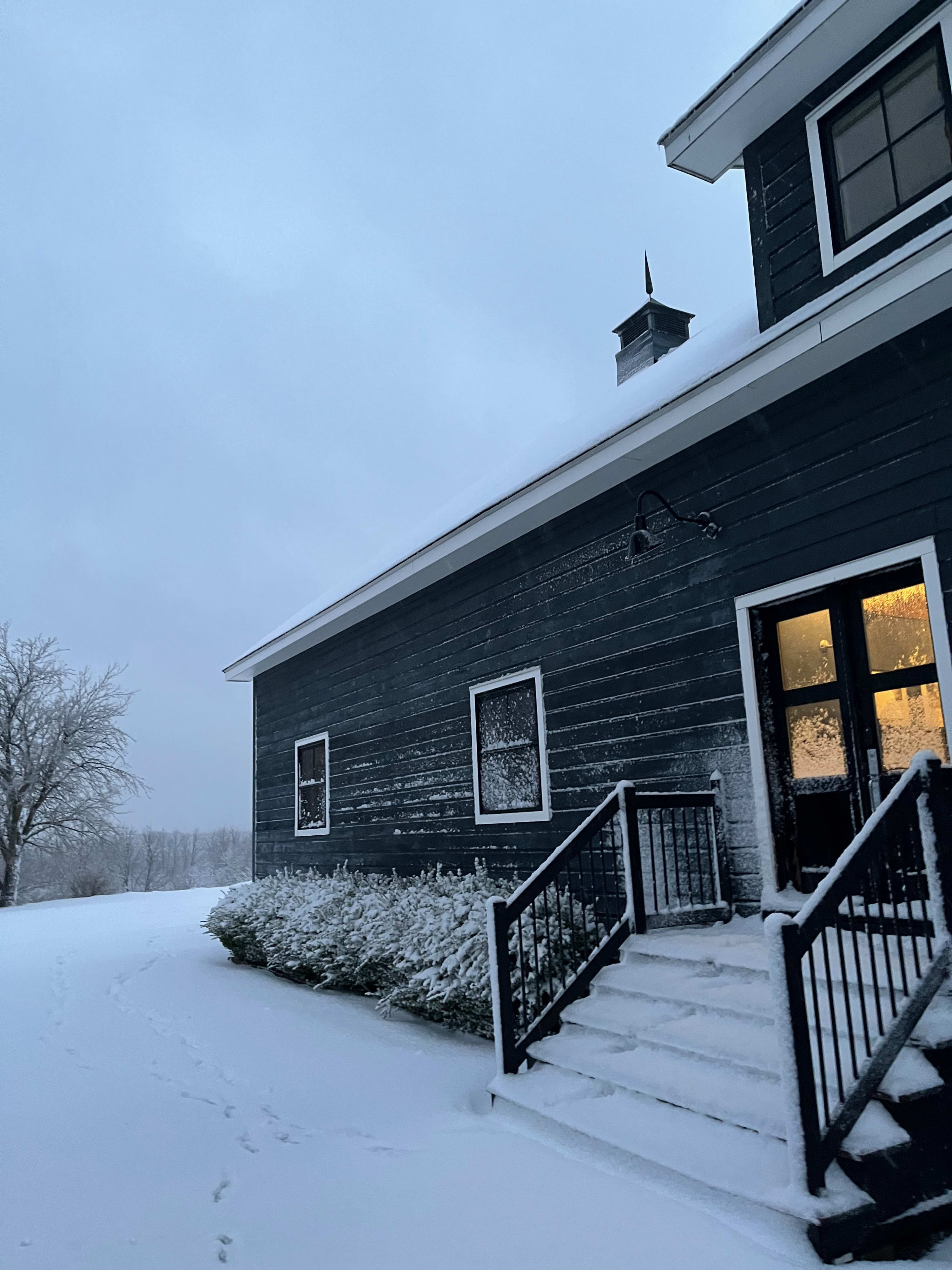 The dark gray barn-style house is covered in a fresh blanket of snow under a cloudy winter sky, with snow-dusted steps leading to the entrance and footprints visible in the foreground.