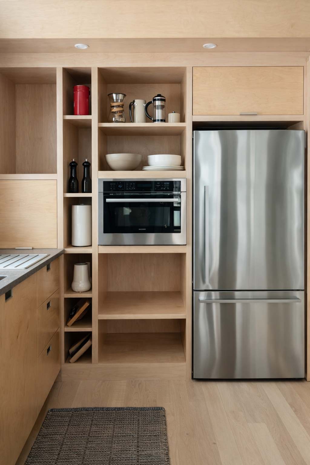 Modern kitchen with light wood cabinetry, stainless steel appliances including a refrigerator and built-in microwave. Open shelving displays kitchenware, and a dark rug sits on the light wood floor.